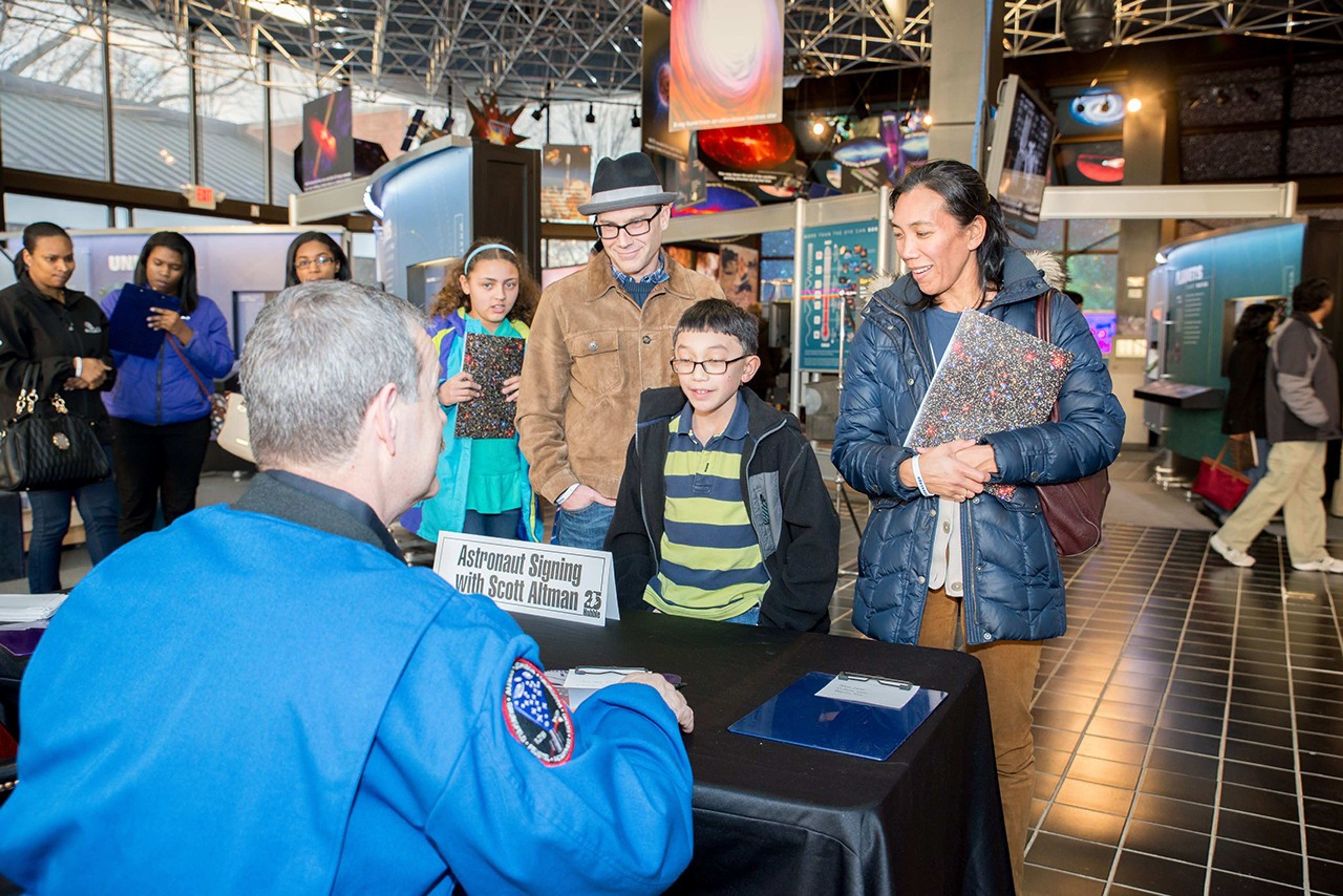Scott Altman sits behind a table and faces away from the camera toward a young boy and the female adult with him, while several other people wait in line on the left side of the image.