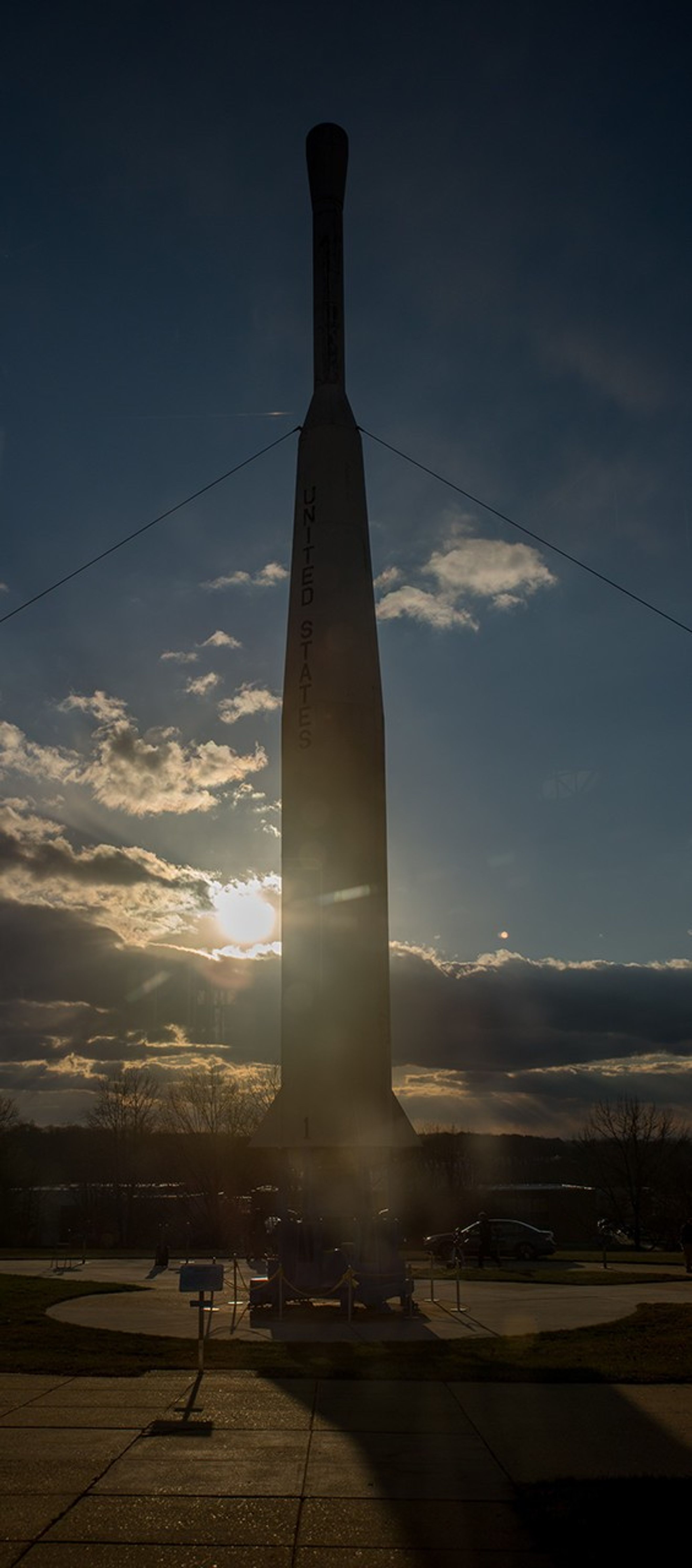 A white Delta-B rocket model with "United States" printed down the side. It is framed in shadow due to the Sun shining from directly behind it to the left from a partly cloudy sky at dusk.
