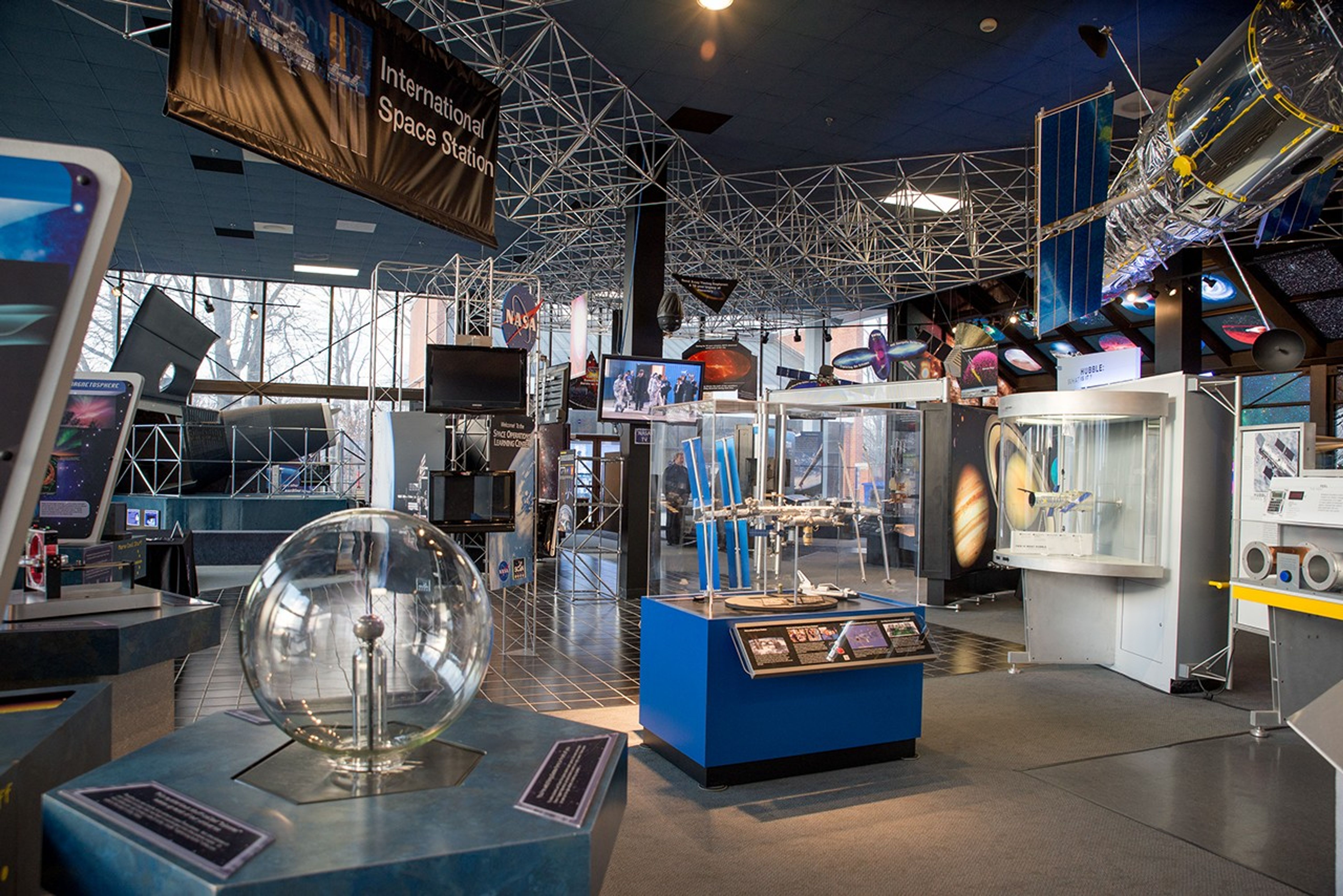 Numerous hands-on activities are visible in the Visitor Center. A model of Hubble hangs from the rafters in the upper-right, along with an International Space Station model in the upper left. In the background, the far wall consists of windows from floor to ceiling that allow visitors to see outside.