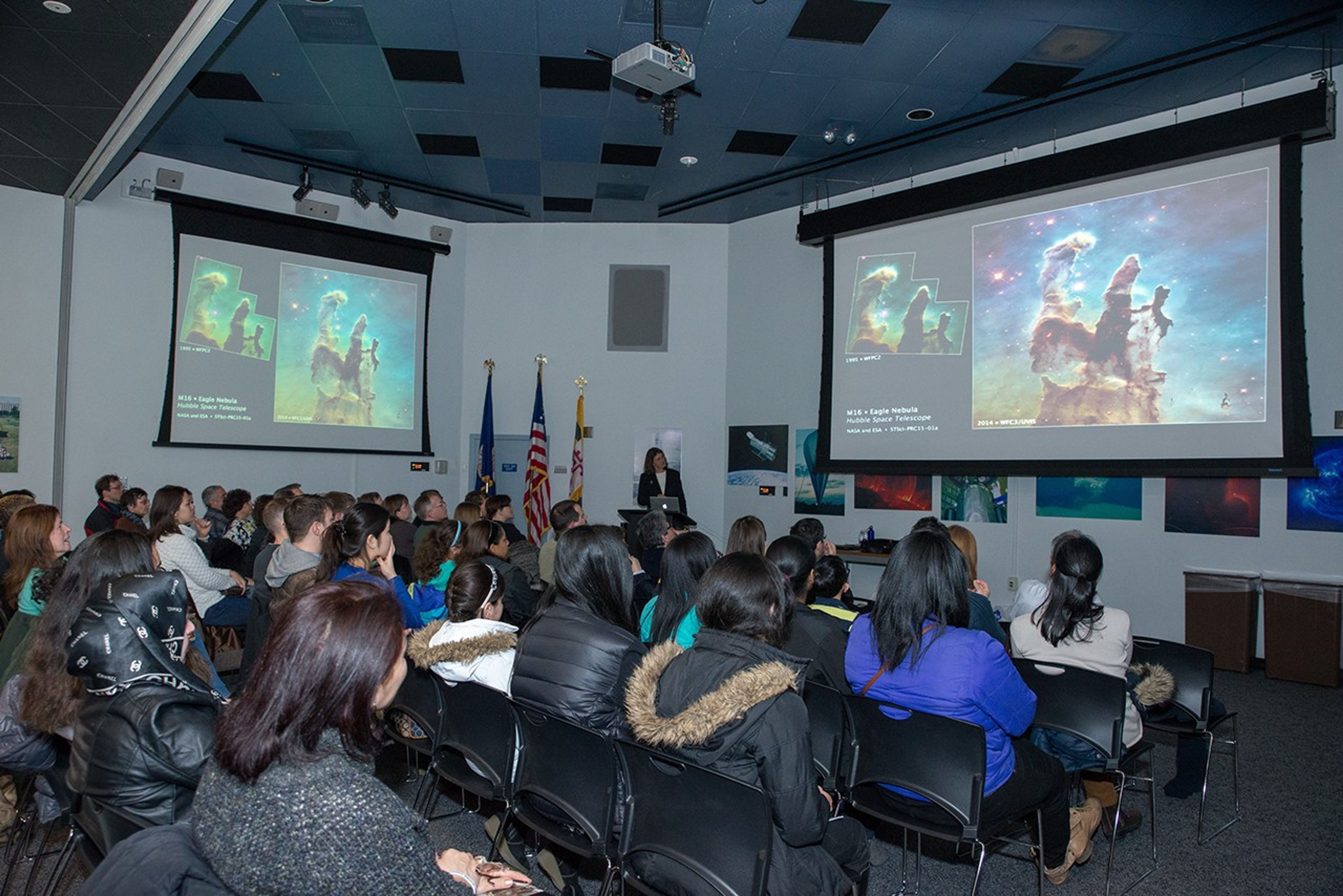 Jennifer Wiseman (in the background on the far side of the room) presents a slide show, projected onto two screens on the right and left, to a large listening audience in the foreground.