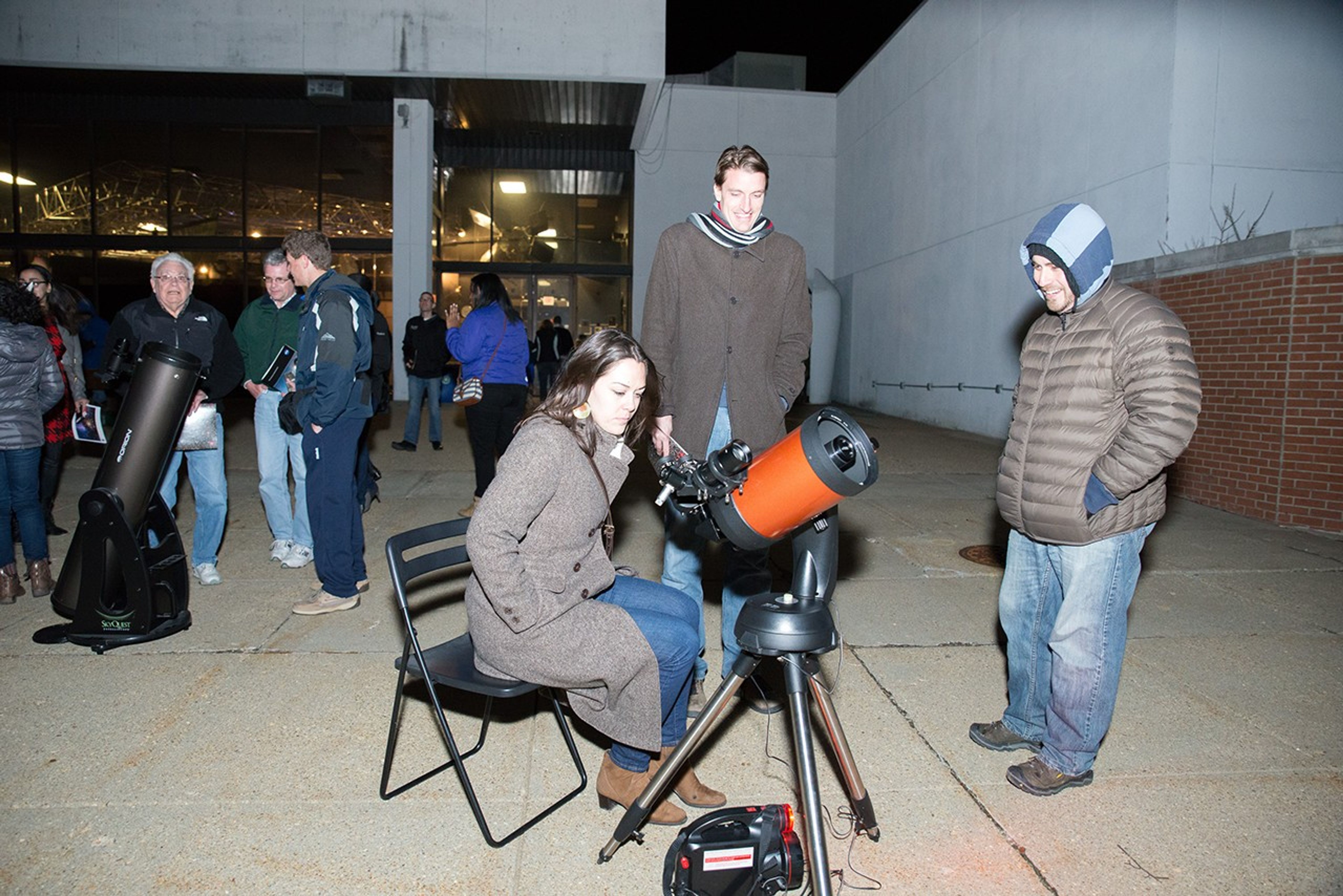 A woman sits in a black chair to look through an orange-and-gray telescope, with two men standing watching nearby. In the background, several more people stand around another dark-gray telescope.