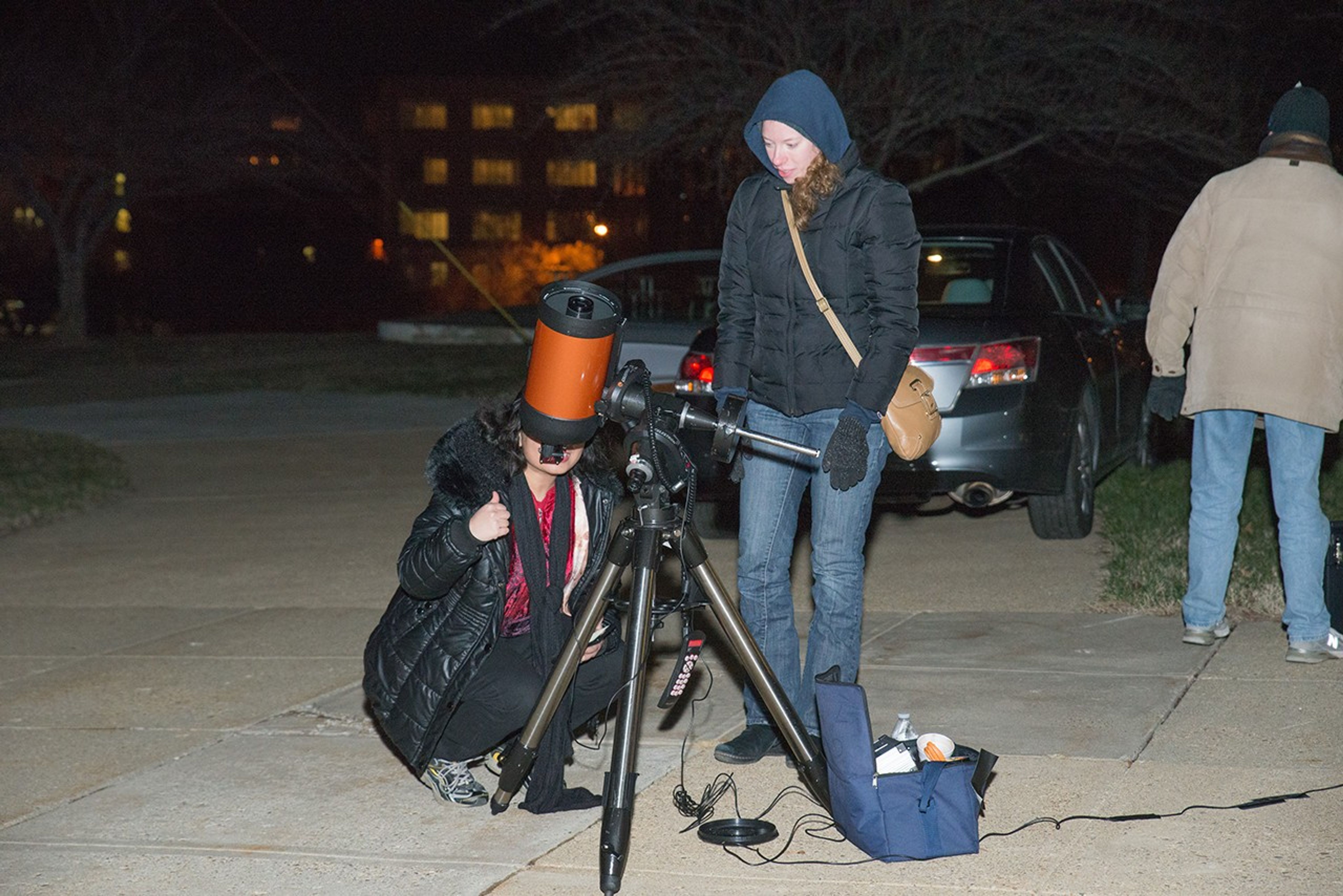 A woman crouches to look through an orange-and-black telescope, while another woman watches.