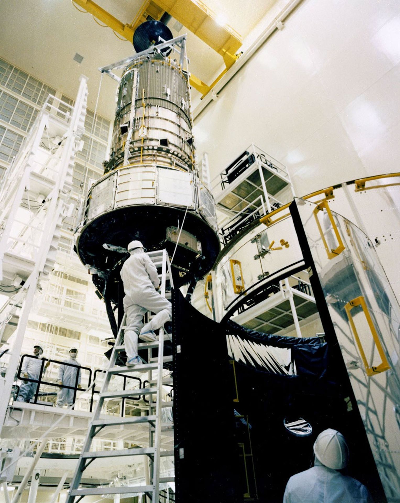 Hubble Space Telescope (HST) being assembled in the clean room of the Lockheed Missile Space Company. Hubble's optical tube and instrument housing appear to be suspended from the ceiling.  Two people, one on a ladder just below Hubble and another on the ground floor are assembling Hubble. Two other workers are on a catwalk to the left of Hubble in this image. All are in protective suits and hardhats.