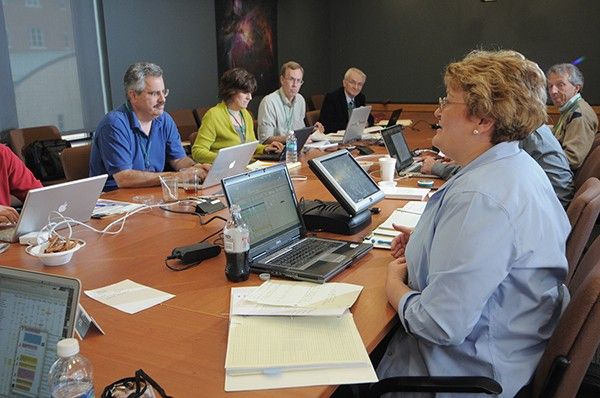 A group of scientists sits at a large conference table, laptops and papers full the table.