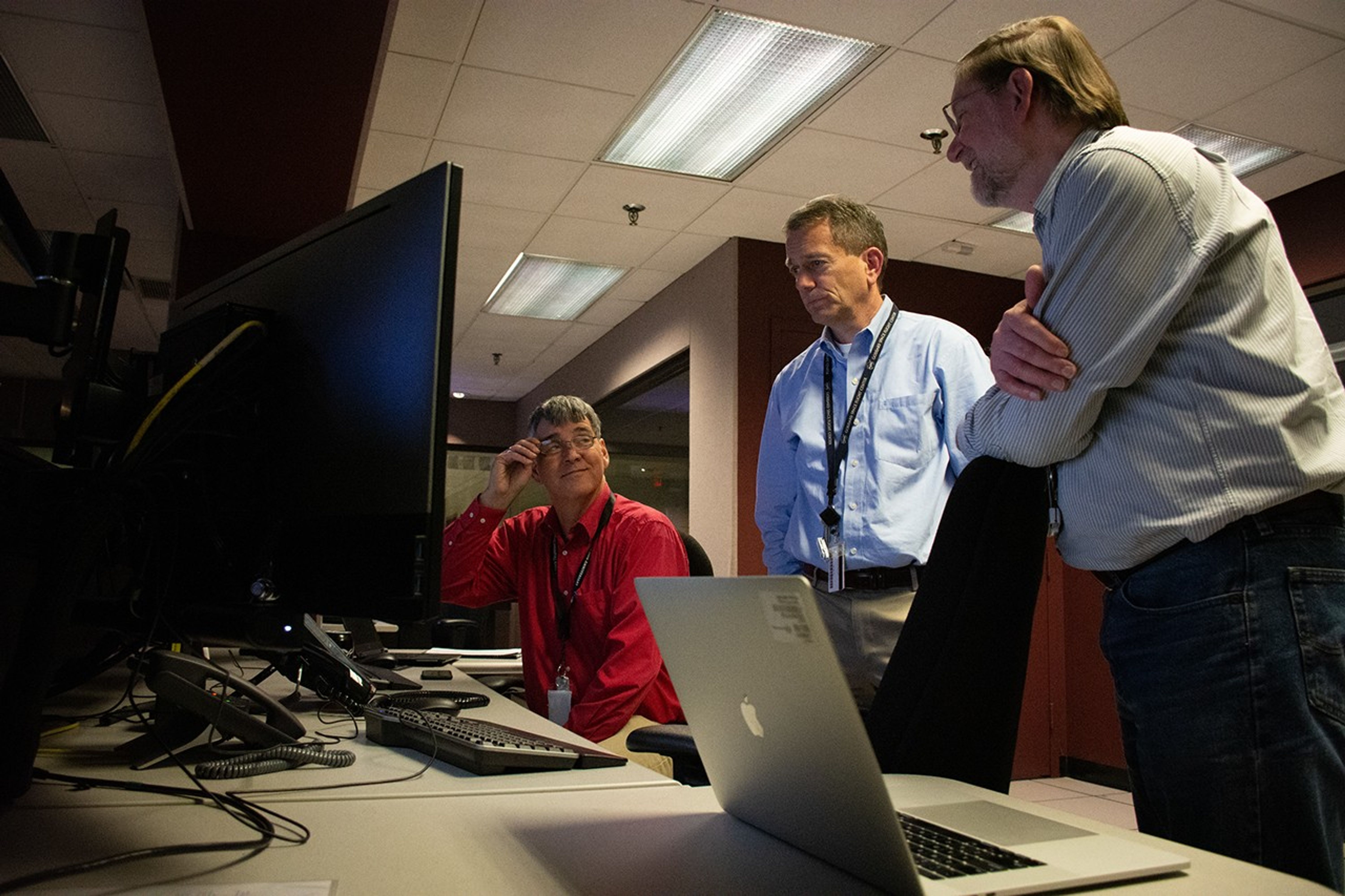 Three men, one sitting and two standing, converse while observing a computer screen. The man in the foreground has his arms folded and is leaning on the back of a chair, the one in the middle has his hands in his pockets, and the seated man is adjusting his glasses.