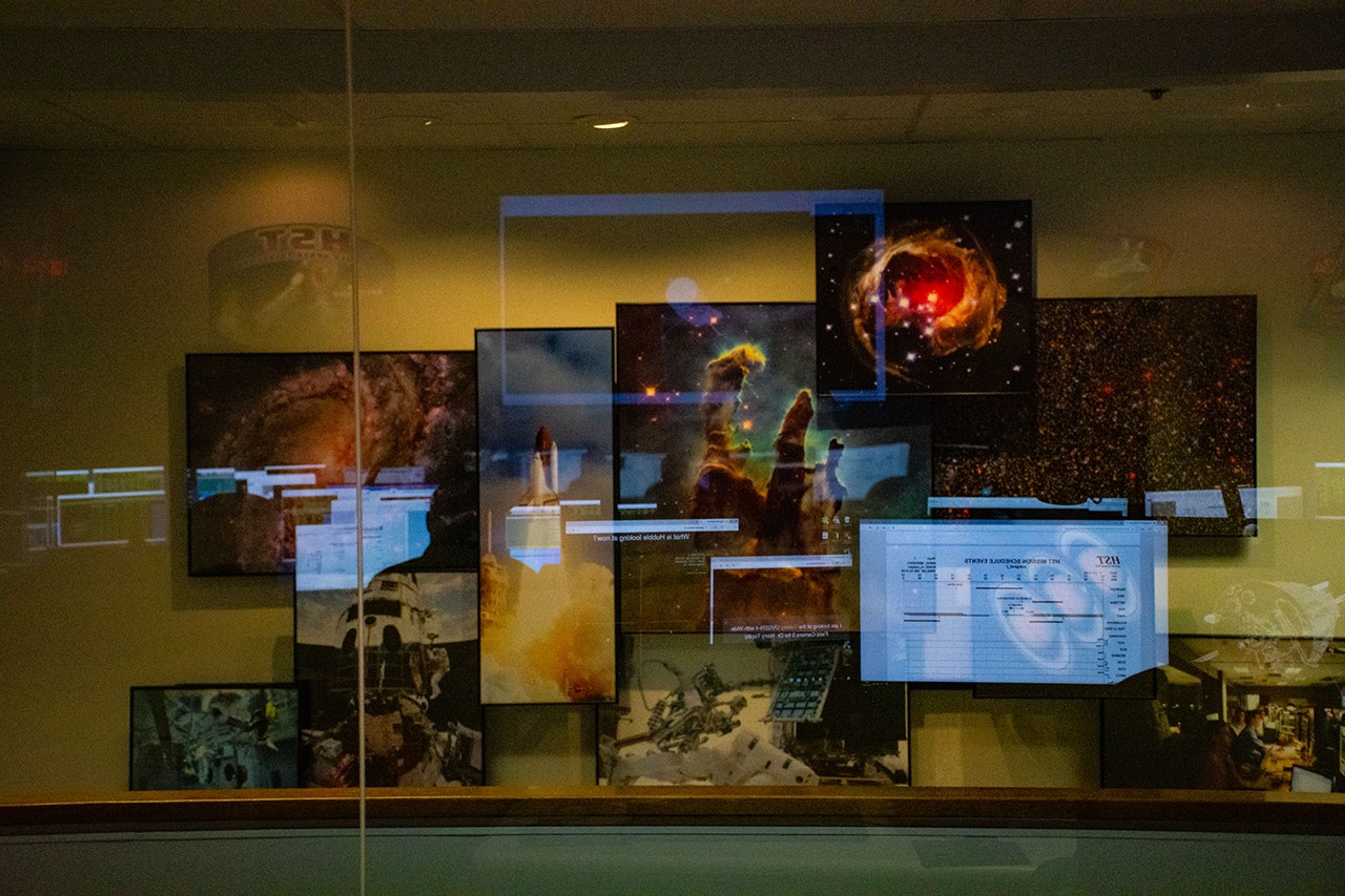 A wall full of Hubble images is viewed through a window. The glow of the computer screens in the room reflects off the window.