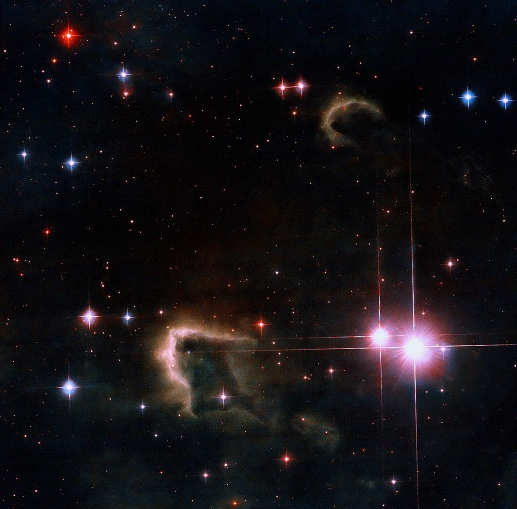 The the light of newly-formed pair of bright pink stars on the right silhouettes two dark, cold, brownish gas clouds in the lower center and upper-right. Many stars of different colors glimmer in the background.