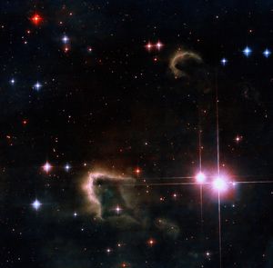The the light of newly-formed pair of bright pink stars on the right silhouettes two dark, cold, brownish gas clouds in the lower center and upper-right. Many stars of different colors glimmer in the background.