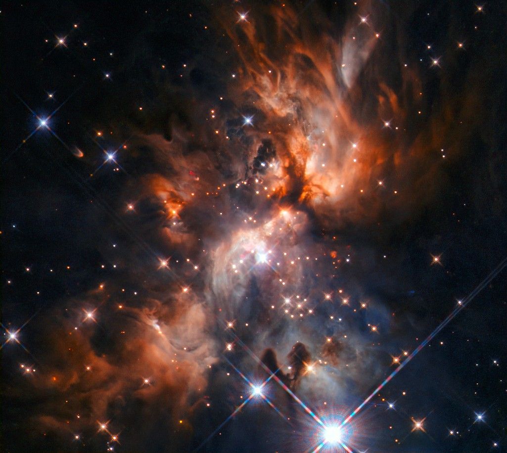 Orange flame-shaped clouds of gas, with dark edges, extend to the upper-left, upper-right, and lower-left of the bright center. A more blue-grayish cloud extends to the lower-right. Bright stars of different colors scattered throughout.