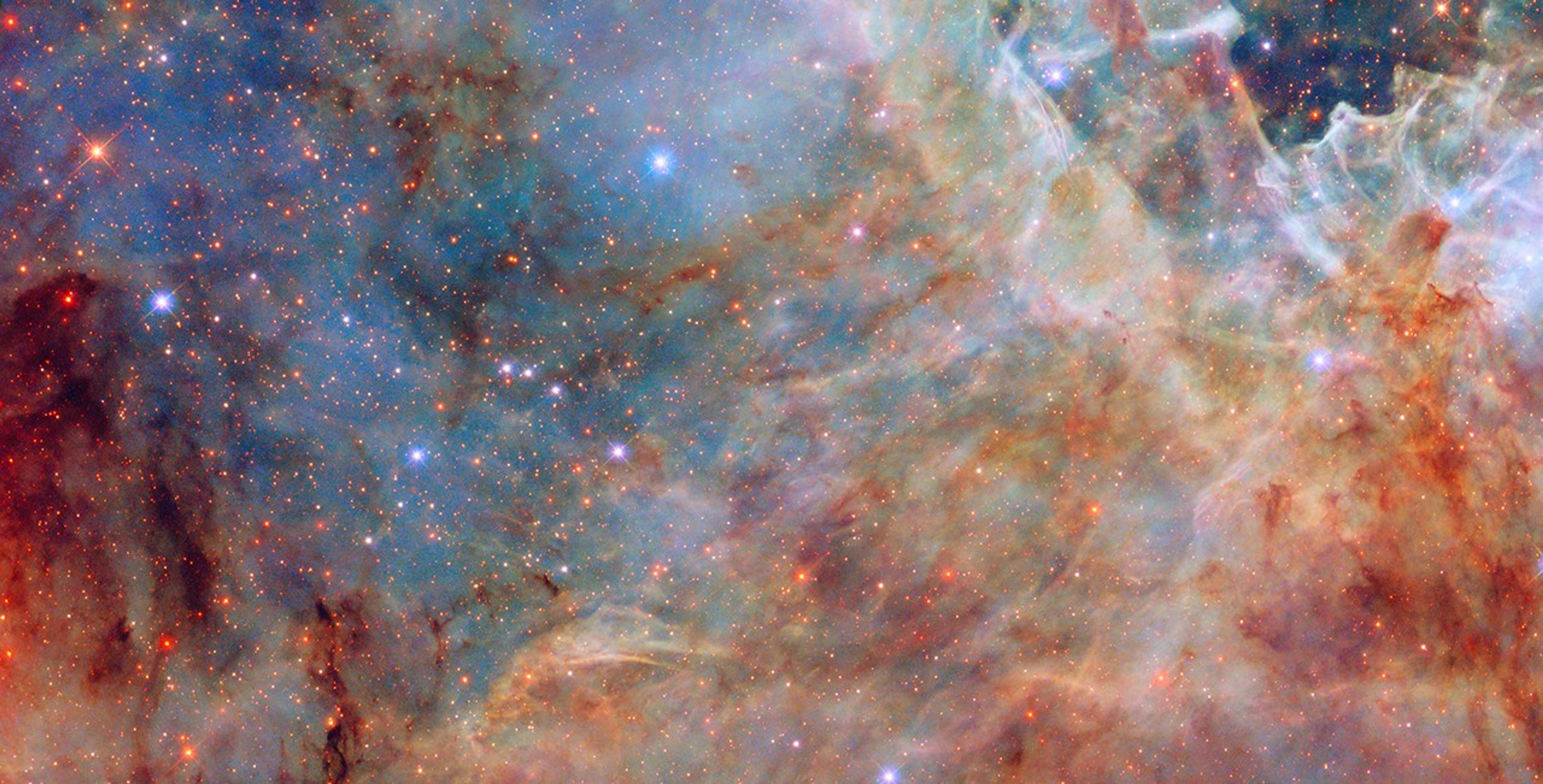A section of Tarantula Nebula that holds layers of colored clouds of gas with varying thickness. In the background are bluish, translucent, and wispy clouds; on top of these are stretches of redder and darker, clumpy dust, mostly along the bottom and right. In the bottom left corner are some dense bars of dust that block light and appear black. Small stars are scattered across the nebula.