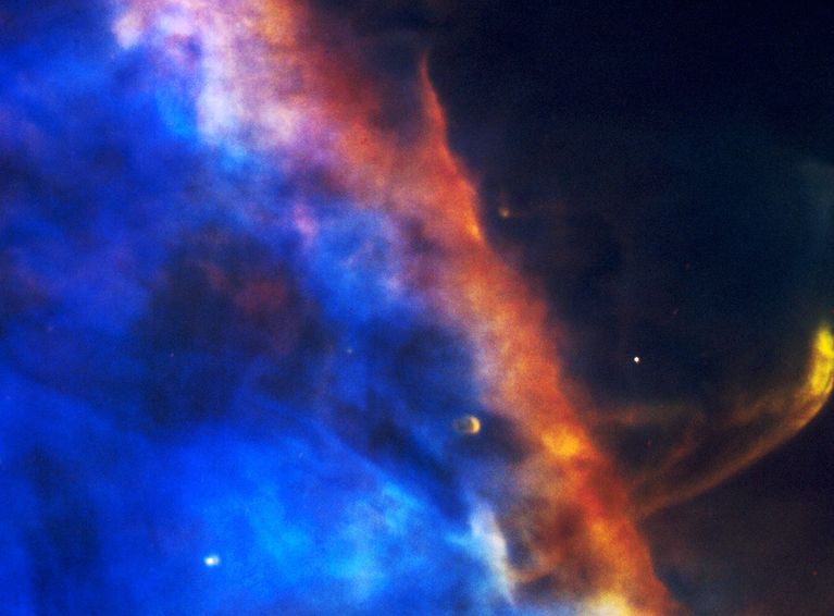 A blue cloud fills the left side of the image.  The blue cloud transitions to white and then reddish yellow toward its edge. A plume of yellow gas extends from the cloud in the lower-right quadrant of the image. one prominent knot in the cloud, near the plume, is a proplyd.