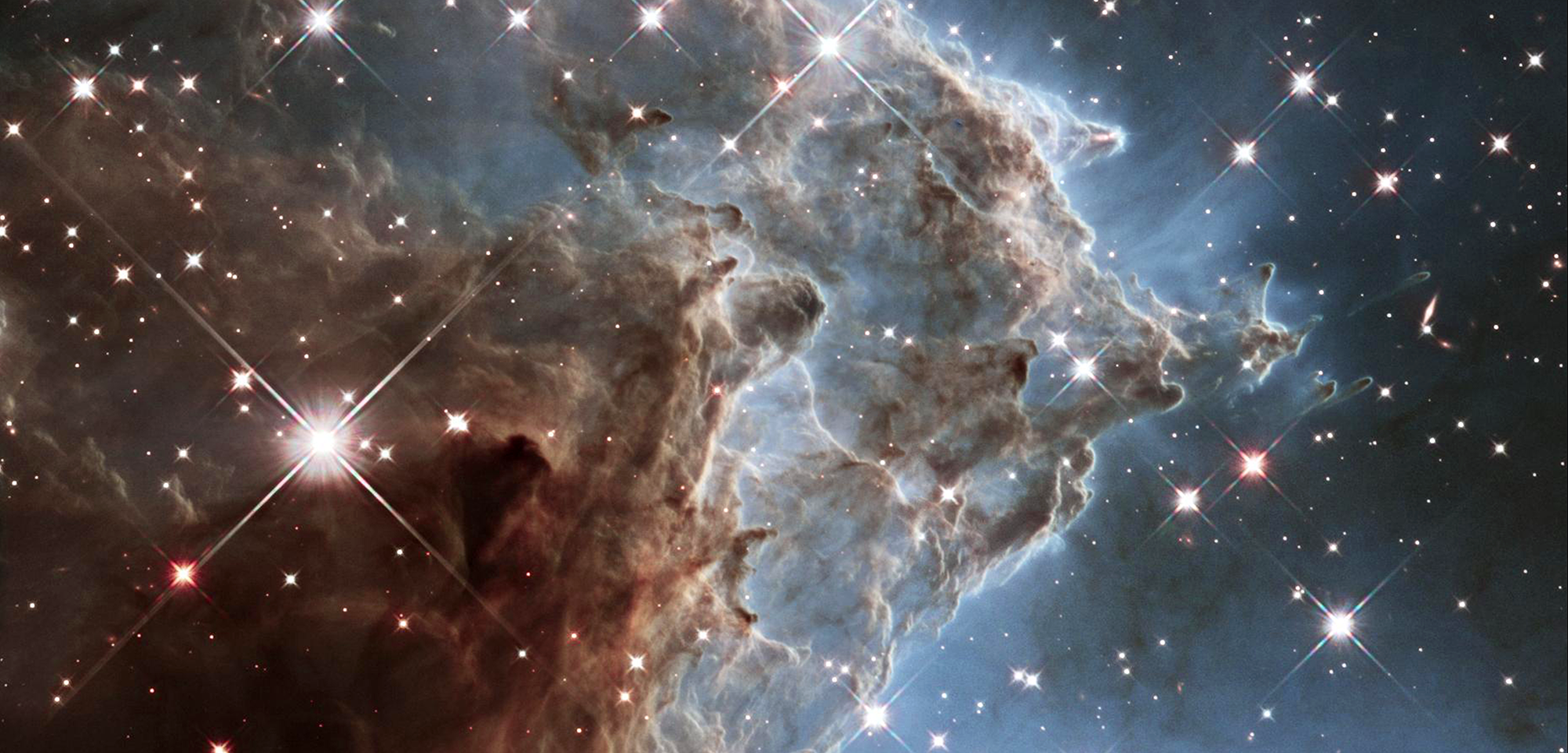 A nebula cloud lightens from left to right, dark brown to light, as it also narrows to form into many peaks. At the edges it is surrounded by a soft blue glow, and many large bright stars shine in its midst, with smaller ones in the far background.