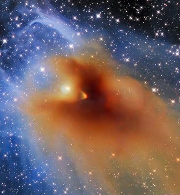 La imagen muestra un objeto naranja brillante de forma irregular compuesto por gas denso y polvo, que parece más oscuro y compacto en el centro. Esta densa nube, llamada CB 130-3, está rodeada por gas y polvo más finos en tonos azules claros. El fondo muestra una multitud de estrellas brillantes sobre un fondo negro.