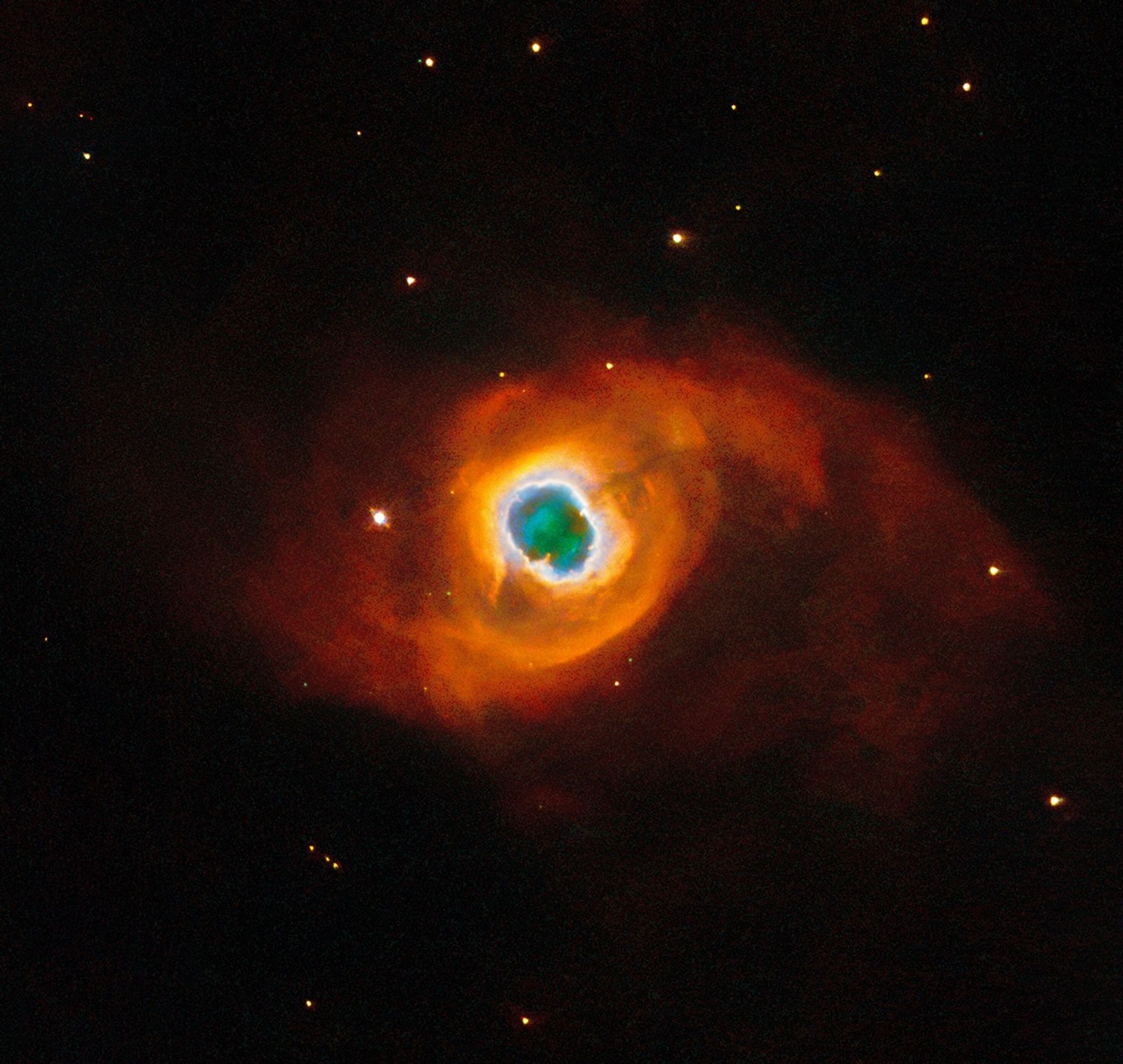The image holds a planetary nebula, a glowing shell of material thrown off by a dying star. A glowing, jagged ring encircles a small central region of greenish clouds, making it appear like a hole torn in fabric. This “hole” is itself encircled by a band of silvery-blue clouds and a larger, fainter, yellow ring of gas. Puffy, smoky clouds of orange and red gas billow out from there into a large oval nebula, fading into the dark background of space dotted by stars.