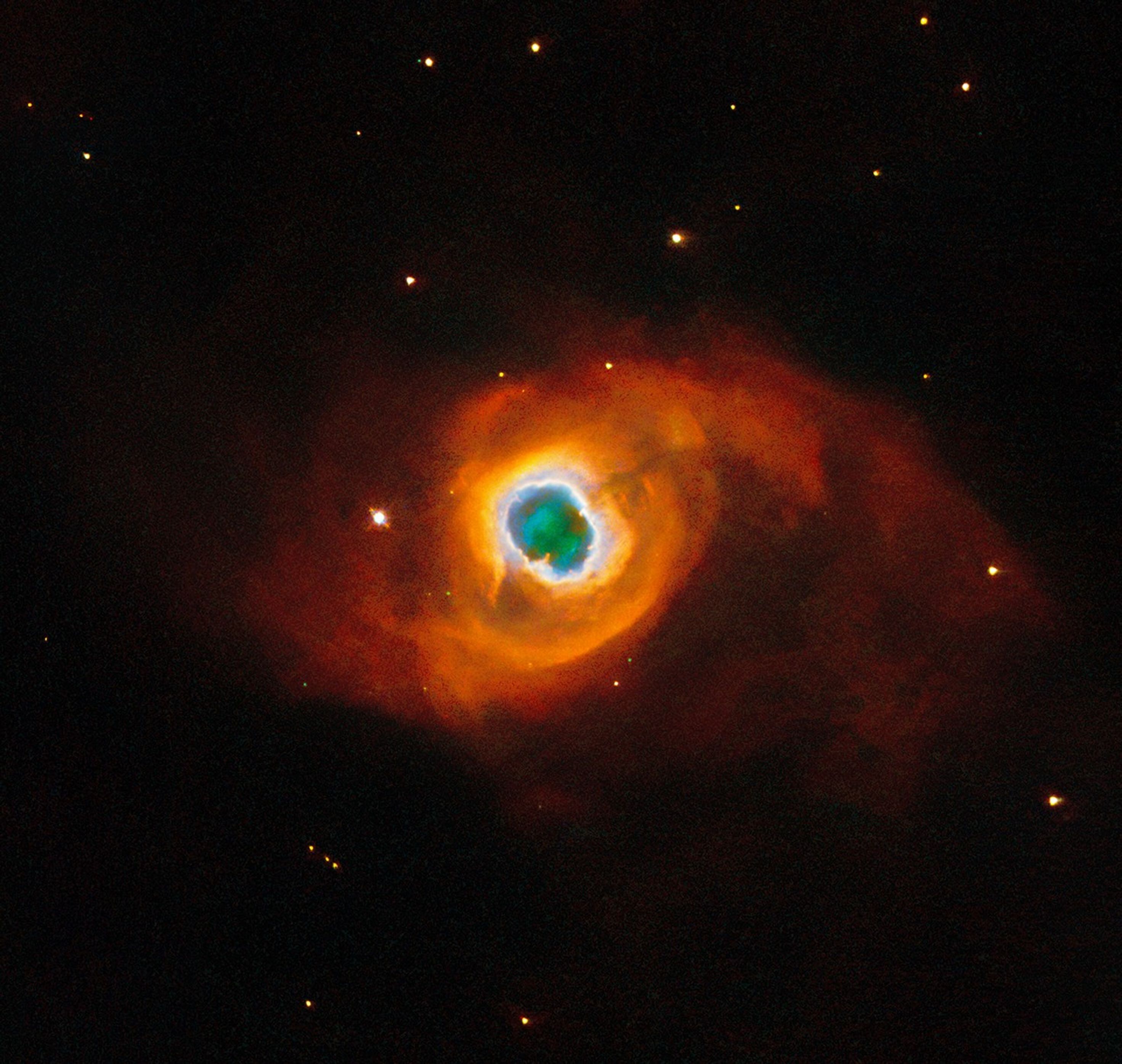 The image holds a planetary nebula, a glowing shell of material thrown off by a dying star. A glowing, jagged ring encircles a small central region of greenish clouds, making it appear like a hole torn in fabric. This “hole” is itself encircled by a band of silvery-blue clouds and a larger, fainter, yellow ring of gas. Puffy, smoky clouds of orange and red gas billow out from there into a large oval nebula, fading into the dark background of space dotted by stars.