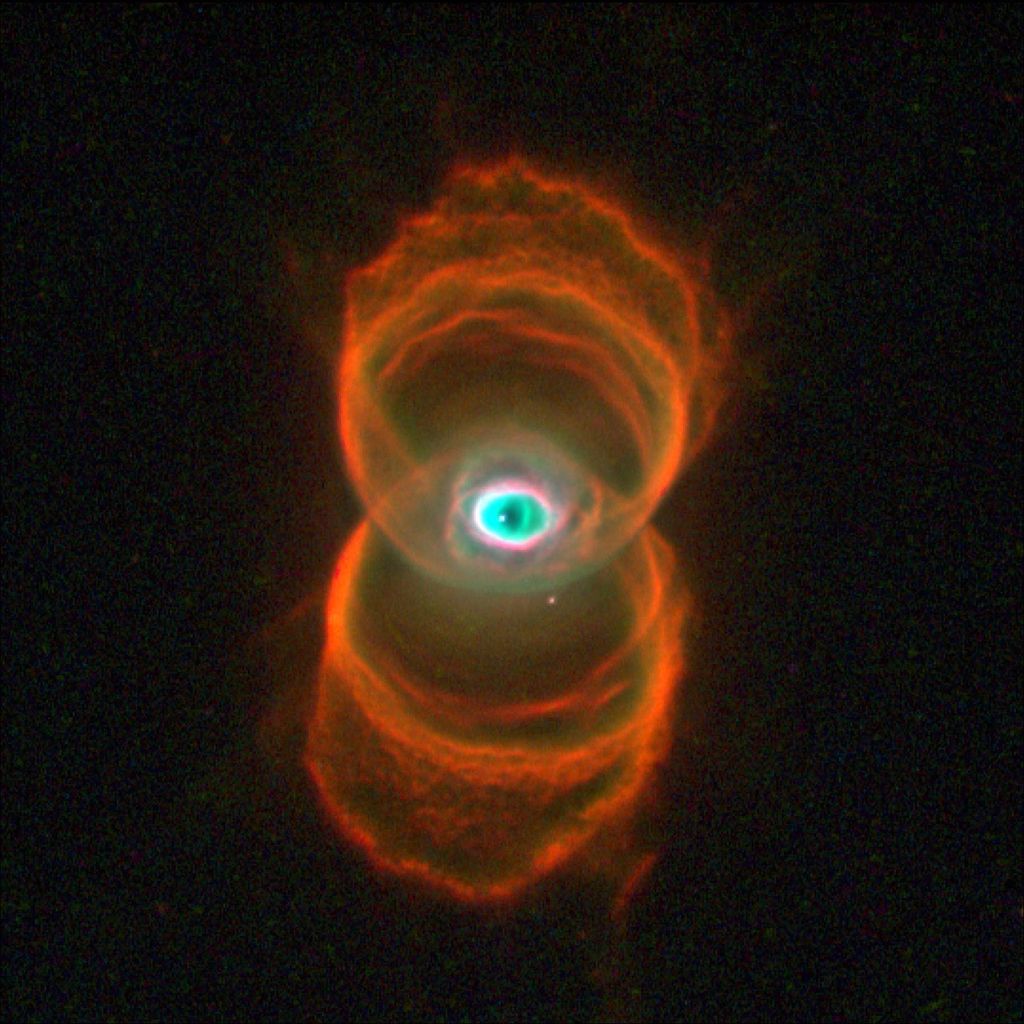 Two sets of rings in orange and gold stacked one above the other form the shape of a hourglass. At image center, where the two sets of rings overlap, is an area of white, green and black dust in a shape that is similar to the human eye.