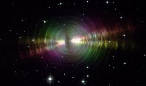 In the image center, an opaque oval cloud of gray gas aligned from 11 o’clock to 5 o’clock hides a star. Two strong beams of light from the star emerge from large holes in both sides of the cloud, forming narrow cones extending toward 8 o’clock and 2 o’clock. The central cloud is surrounded by concentric, wispy shells of gas illuminated by the star’s light. The shells reflect extra light where they’re hit by the twin beams. These clouds and beams appear in a rainbow of colors. A crowd of smaller stars with cross-shaped spikes over them surrounds the nebula on a black background.