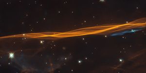 A long, thin, twisted ribbon of orange gas and dust stretches from left to right across the image. Bright-white stars dot the black background. One bright, blue-white star at bottom left. A small swath of blue gas stretches below the orange ribbon on the right side.