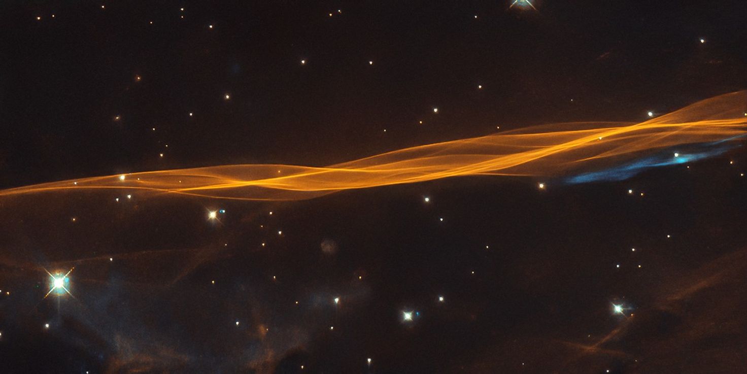 A long, thin, twisted ribbon of orange gas and dust stretches from left to right across the image. Bright-white stars dot the black background. One bright, blue-white star at bottom left. A small swath of blue gas stretches below the orange ribbon on the right side.