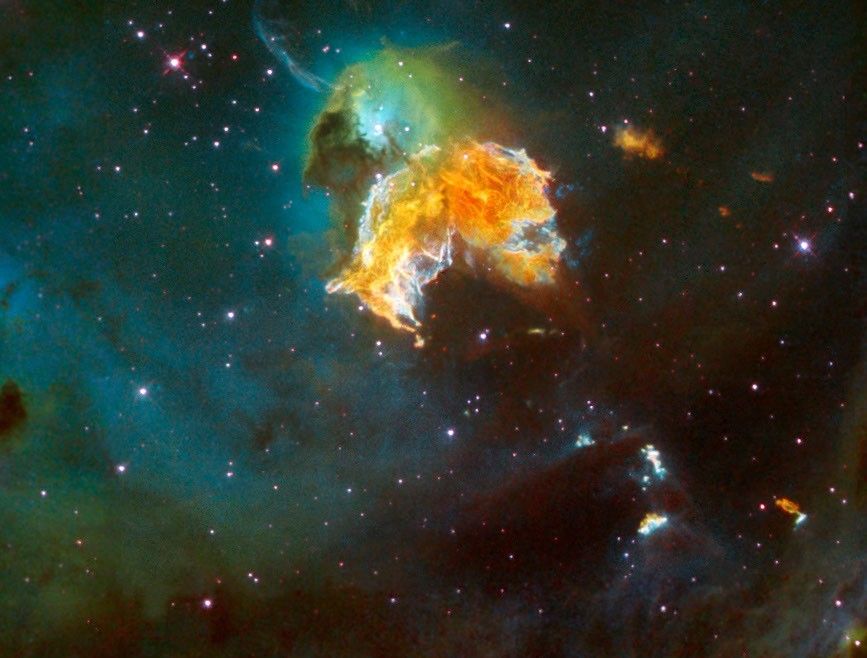 A yellow-orange flair of gas, surrounded by white, shaped like an angel's wings. Above it is a green cloud of dust surrounding a blue haze around a star at the center. It's surrounded by blue haze, with dark green gas at the edges and many stars in the background.
