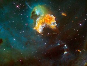 A yellow-orange flair of gas, surrounded by white, shaped like an angel's wings. Above it is a green cloud of dust surrounding a blue haze around a star at the center. It's surrounded by blue haze, with dark green gas at the edges and many stars in the background.