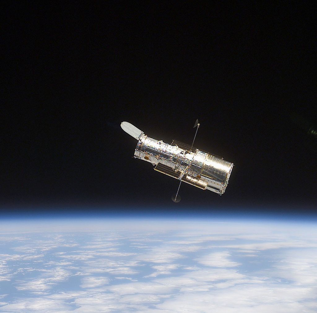Hubble is visible against the blackness of space at the center of the image. The telescope is facing and is pointing in the direction of the upper-left corner of the image. Earths blue limb is visible on the lower third of the image. The planet's blue oceans and puffy-white clouds are visible.
