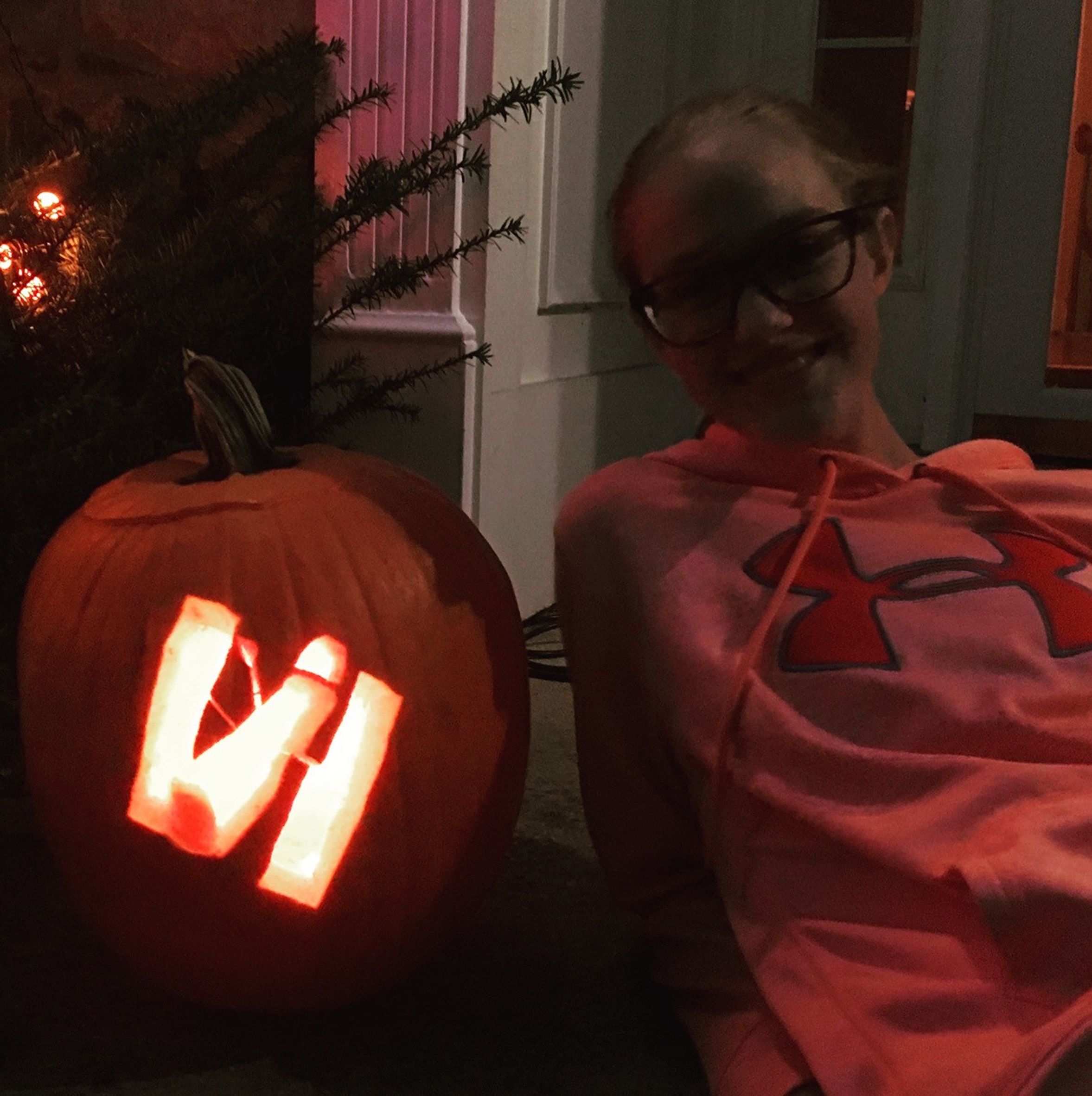 A girl in an orange/pink sweater, right, sits on a porch in faint evening lighting. Next to her, left, is a jack-o-lantern with the shape of the Hubble Space Telescope carved into it.
