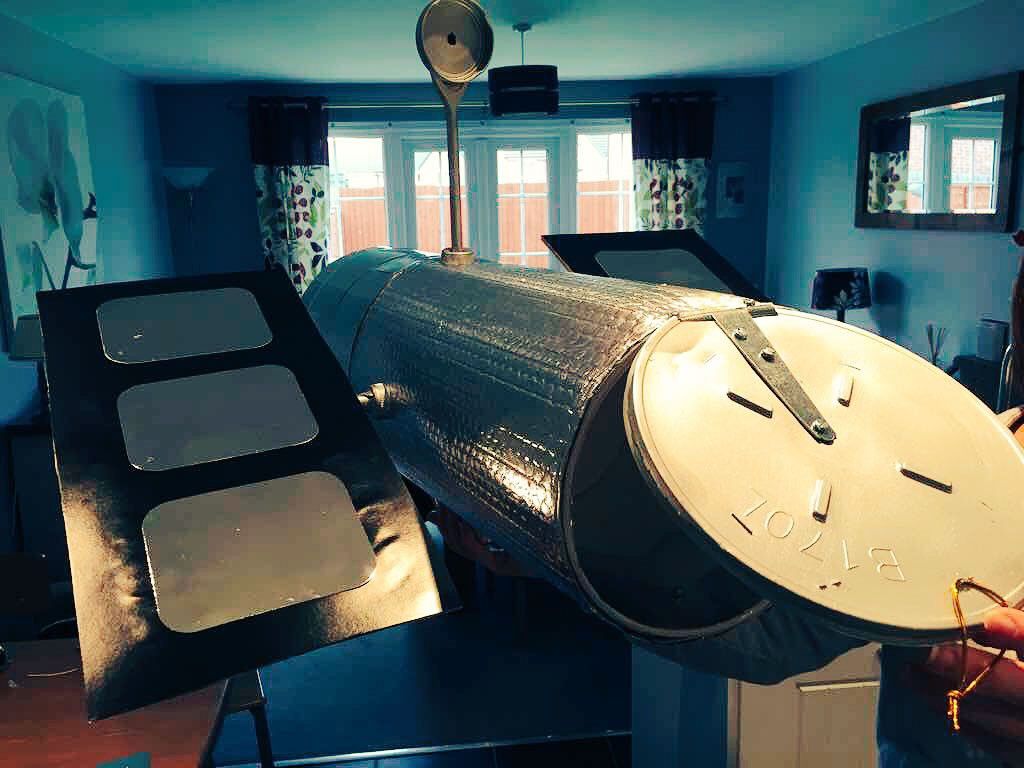 In someone's living room (which is visible in the background) sits a large model of the Hubble Space Telescope, built with home materials like a metal trash can and lid.
