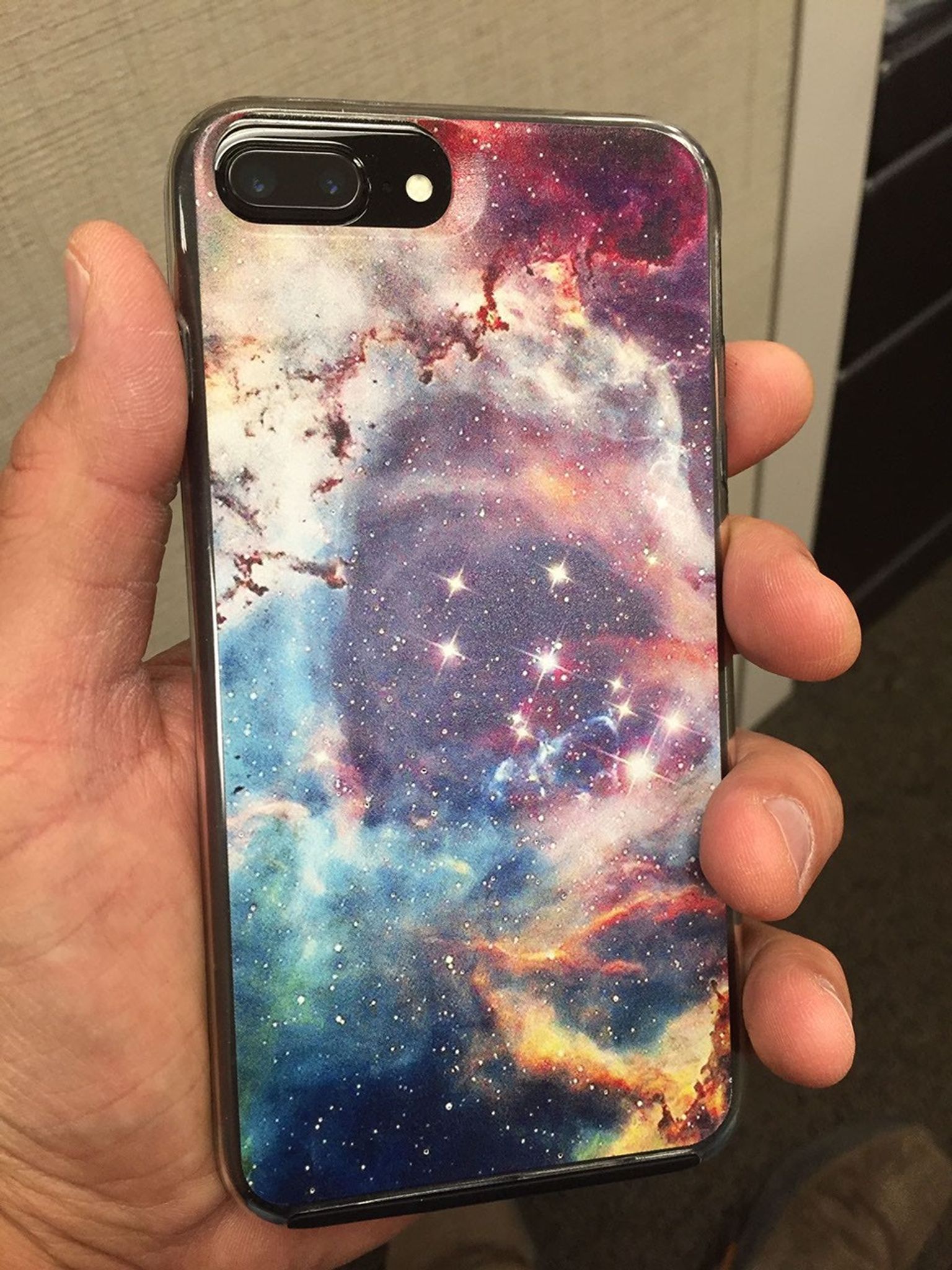 In their left hand, a person holds a smart phone facing away from the camera. The back of the phone's cover has a beautiful image of a colorful nebula with a smattering of bright stars in the center.