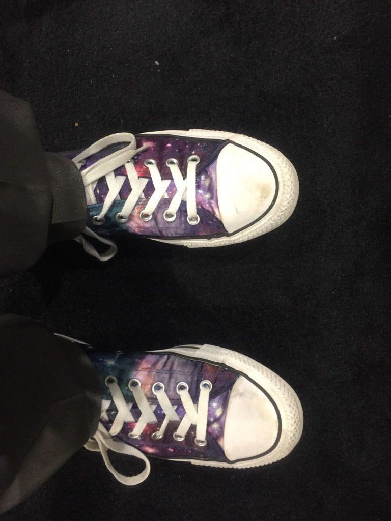 A picture of shoes someone is wearing, with the bottoms of their black pants visible. The sides, toes, and shoelaces of the shoe are white, but the design patter of the top of the shoe is space-themed, showing the purples, pinks, blues, and whites of a Hubble nebula.