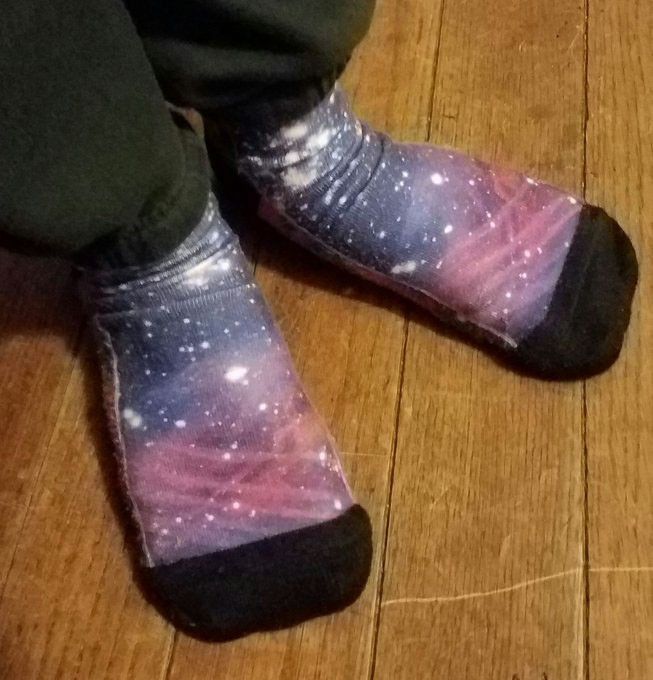 A person wears socks with black toes, and the dark gray bottoms of their pants are visible. The space patterns on the socks include nebulae of pink, light purple, and dark blue, with white dots for stars.