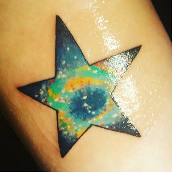 A star-shaped tattoo on a person's arm. The inside of the star is designed to look like the Helix Nebula, with shades of mostly blue and green, with swirls of yellow and white dots to represent stars.