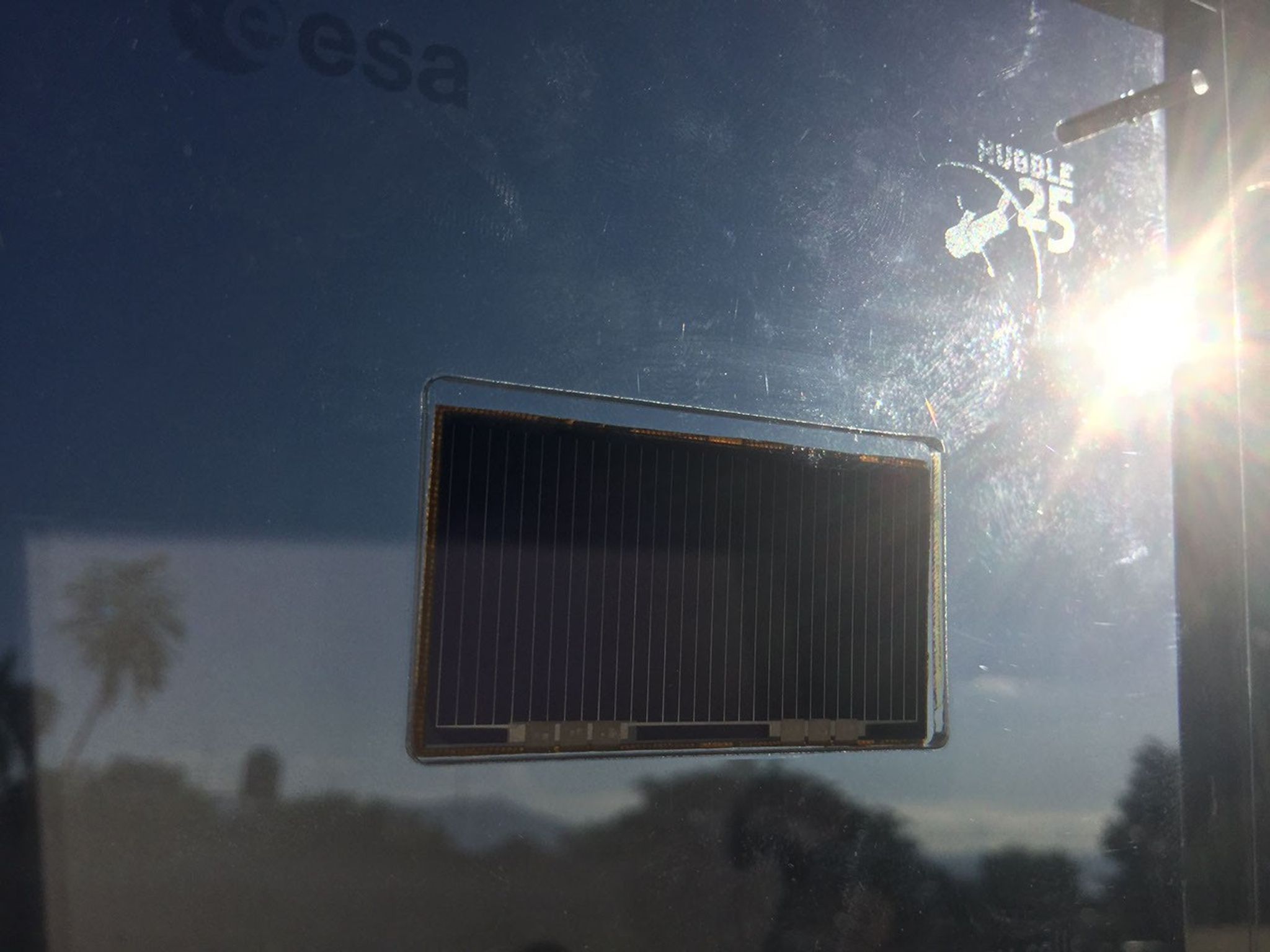A reflective panel/wall with a rectangular solar panel in the center. To the upper right of it is a monogrammed "Hubble 25" and a logo of Hubble, and to the upper left, the logo of ESA. Sunlight reflects off the wall.