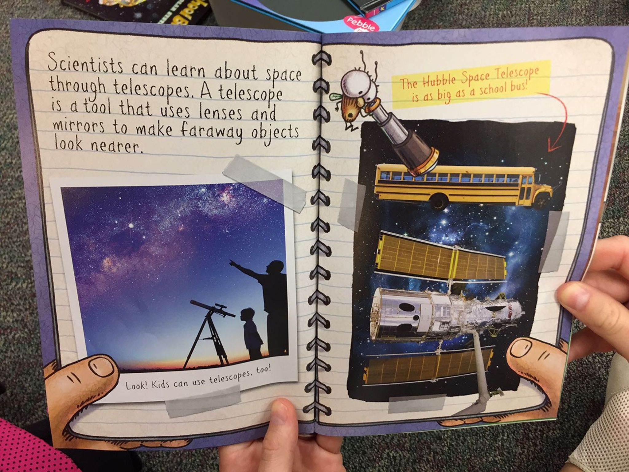 A person holds a children's book in both hands. The left page contains words and a picture of an adult and child using a backyard telescope. On the right page is a graphic showing the Hubble Space Telescope with a space background and a school bus above it, showing that the sizes of the two are comparable.