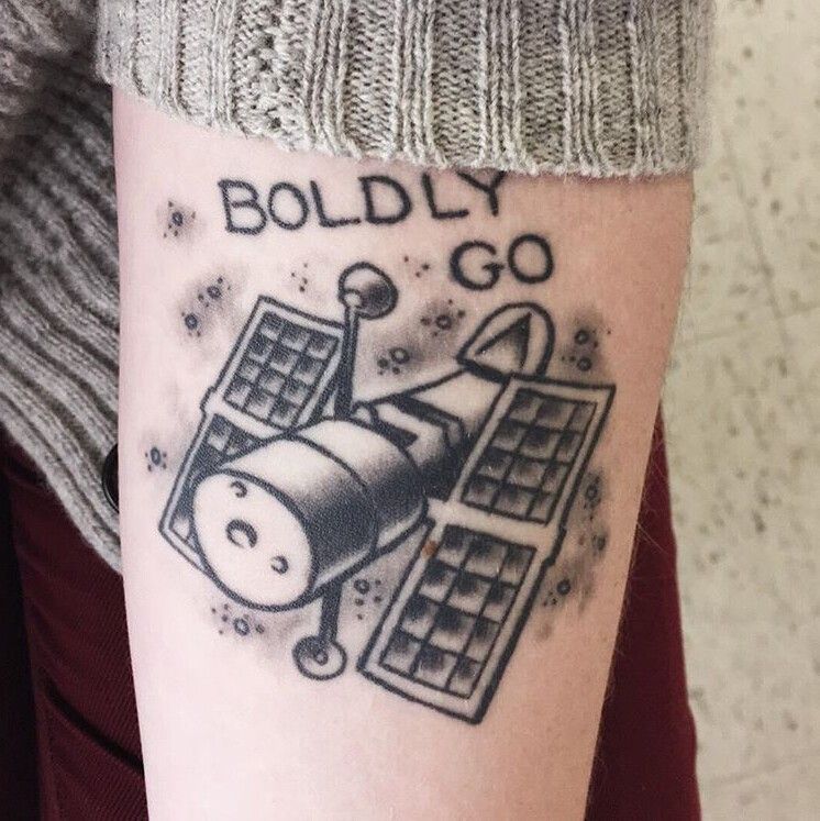 A person has a black-ink tattoo of the Hubble Space Telescope on their arm, with the words "BOLDLY GO" above it.