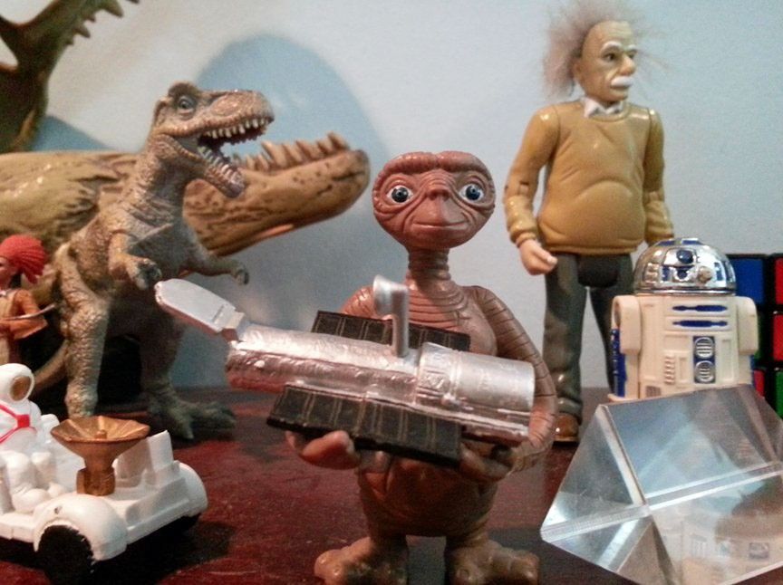 In a group of plastic toys on a shelf including dinosaurs, a Mars rover (both left), Albert Einstein, R2-D2, and a glass prism (all right), a plastic alien toy in the center holds a plastic model of the Hubble Space Telescope.