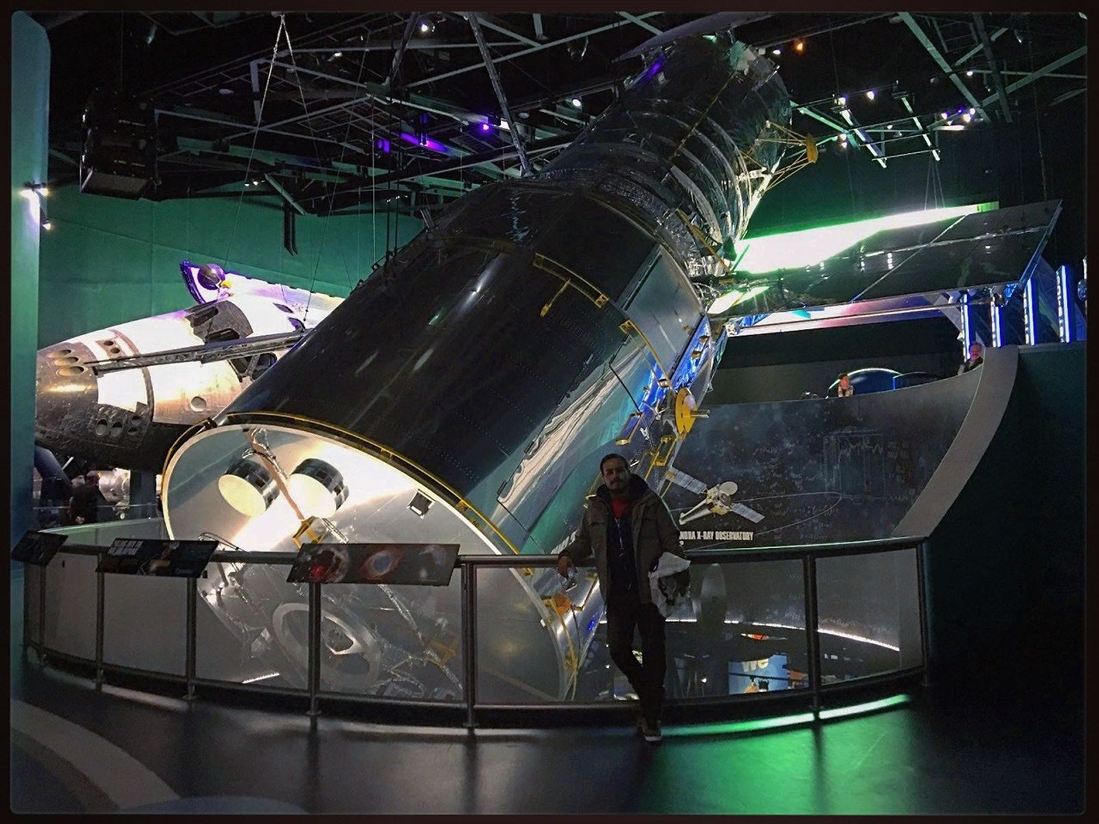 A man poses for the camera in front of a railing, which has panels with Hubble images on it. Behind him, suspended from the ceiling, is a life-size model of the Hubble Space Telescope.