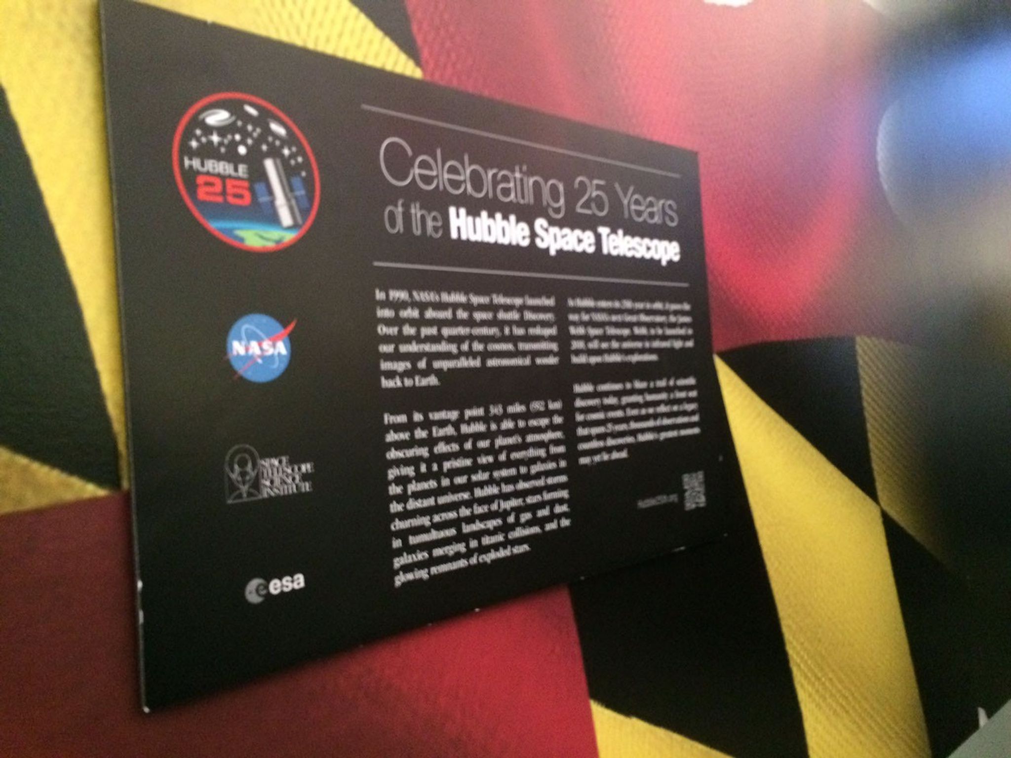 A panel is mounted on the wall that has a column of logos on the left: the Hubble 25th anniversary logo at the top, then the NASA, Space Telescope Science Institute, and ESA logos below it. On the right, there's a header that says "Celebrating 25 Years of the Hubble Space Telescope", with more text underneath it. It's mounted on a wall that's painted to look like the Maryland state flag.