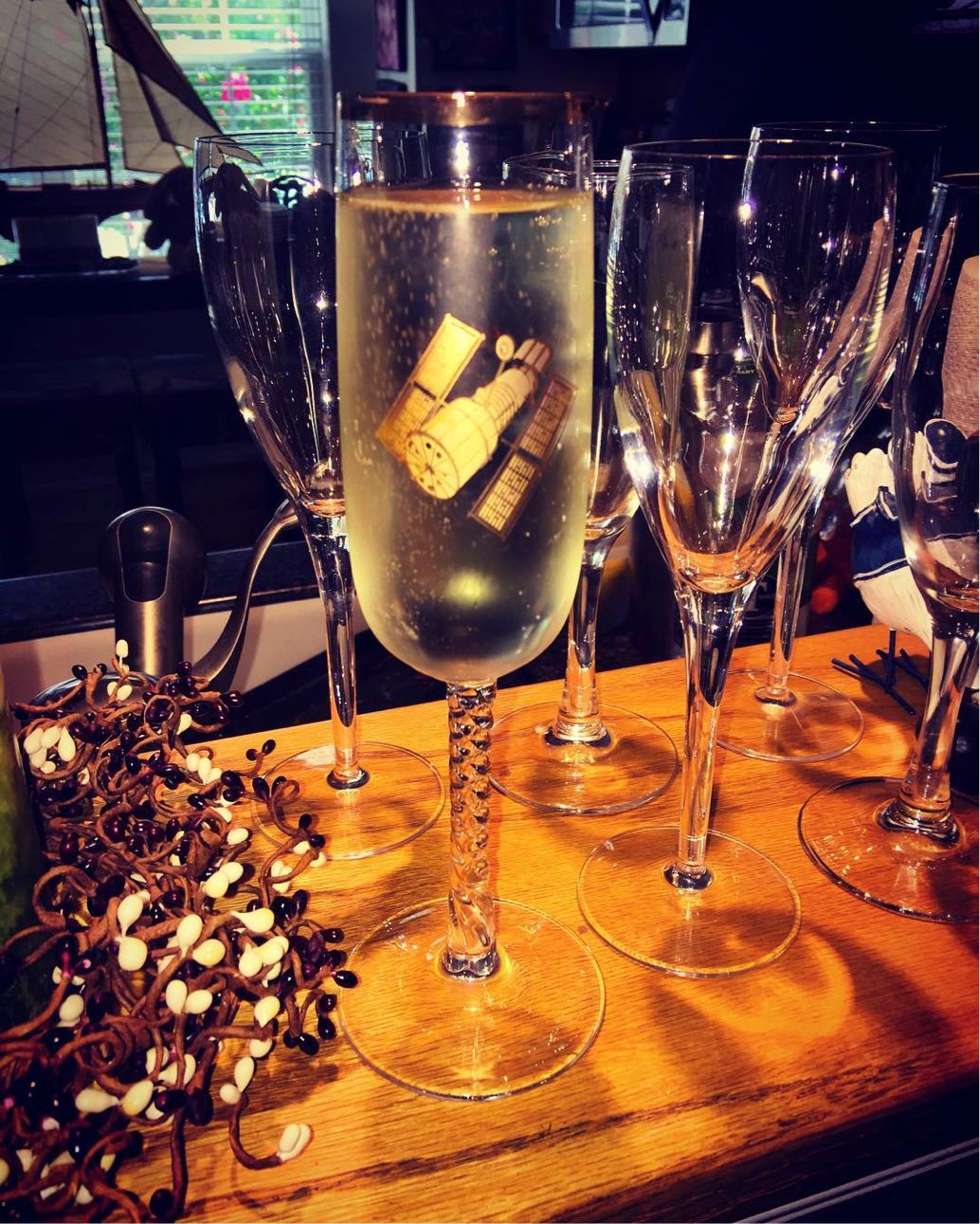 A series of two rows and three columns of champagne glasses sit on a table. The one in the foreground of the image, which is full of champagne, has a monogram of the Hubble Space Telescope on it.