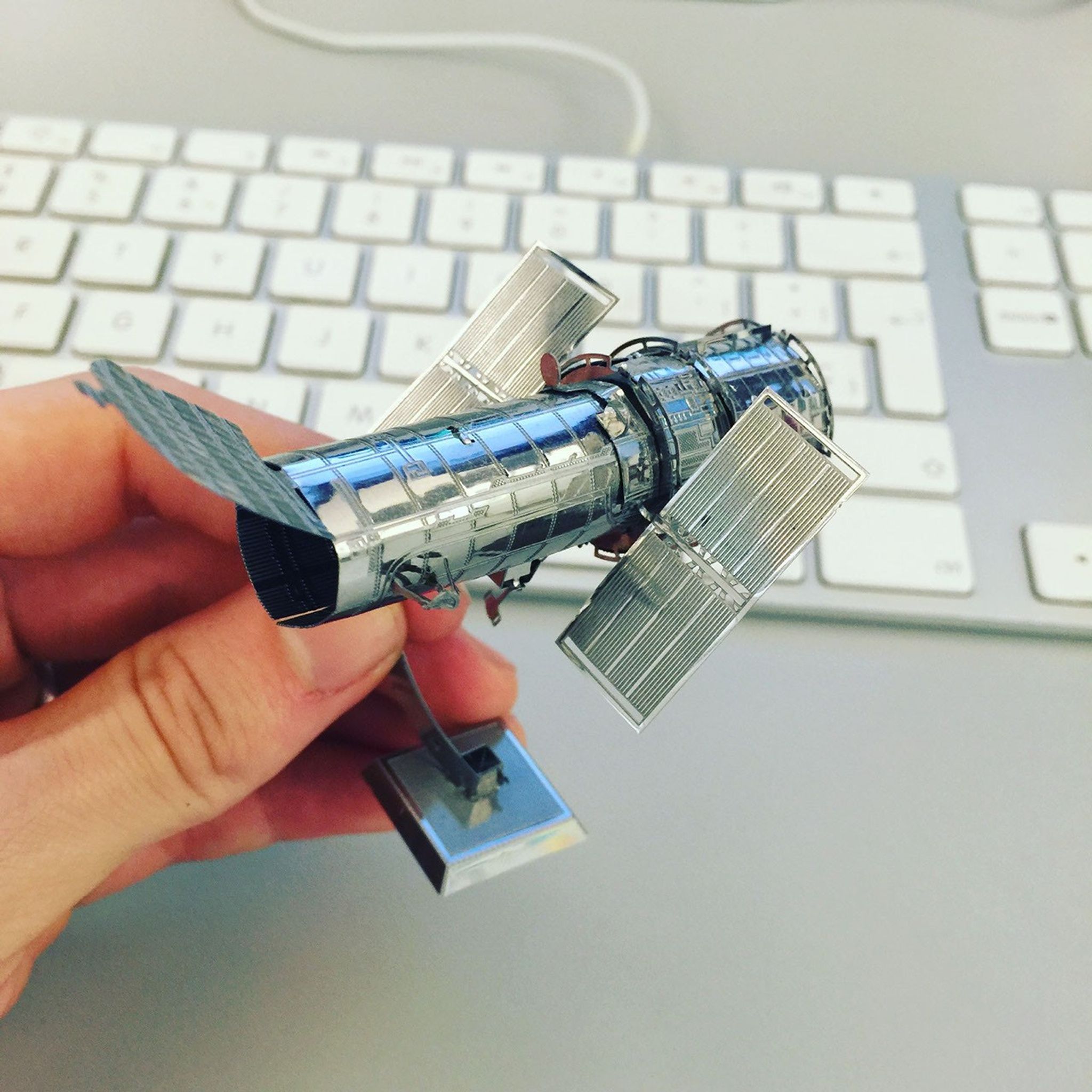 A person holds a small metal model of the Hubble Space Telescope, with an Apple keyboard in the background.