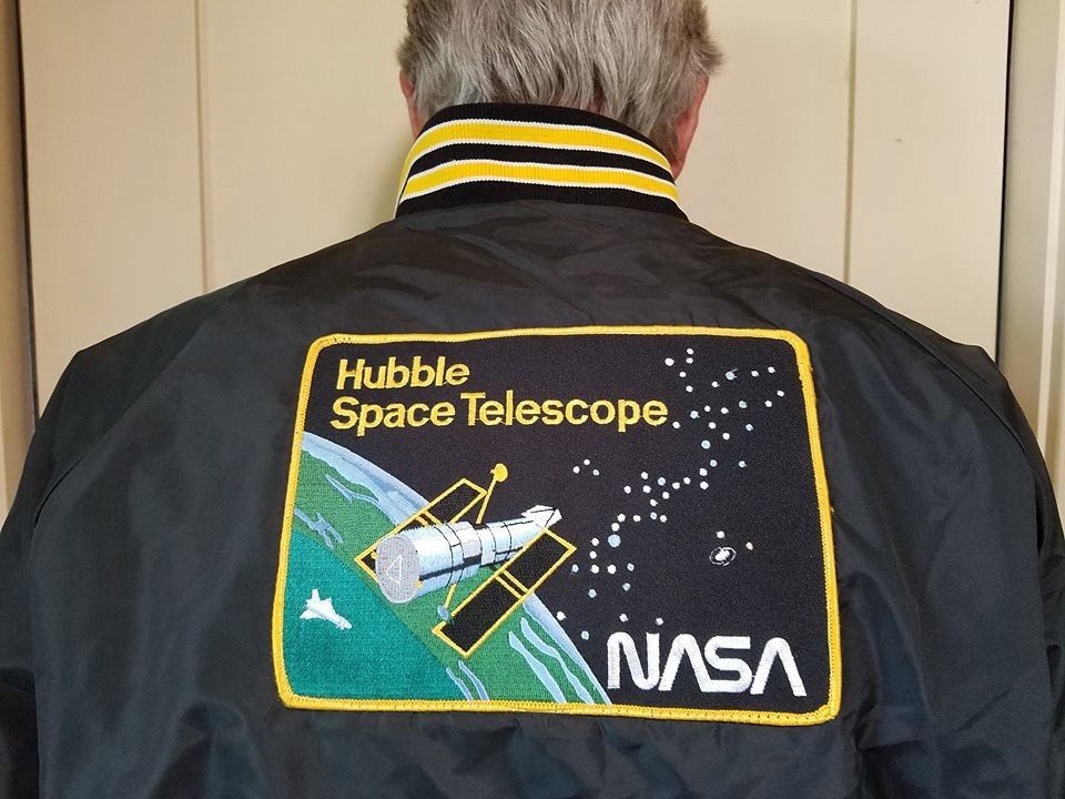 A man faces away from the camera. On the back of his jacket is a large patch with a yellow boarder that says "Hubble Space Telescope" in yellow in the upper-left and "NASA" in white in the lower-right. The picture on the patch shows Hubble over the Earth, pointing out at the stars and galaxies in space.