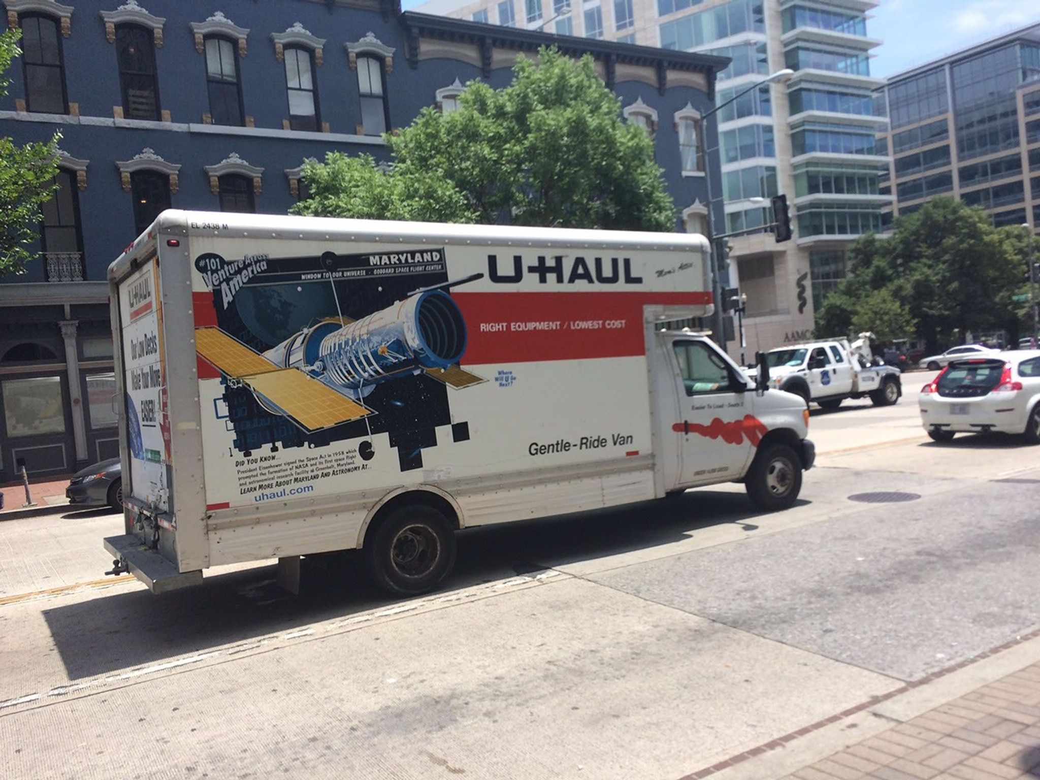 A U-Haul truck, facing toward the right side of the image, is parked b a curb on a sunny day, with buildings and other cars in the background. On the right side of the truck is a square-ish picture that says "#101 Venture Across America" in the upper-left, "MARYLAND" in the upper right, and below them, a large digital image of the Hubble Space Telescope against a space background.