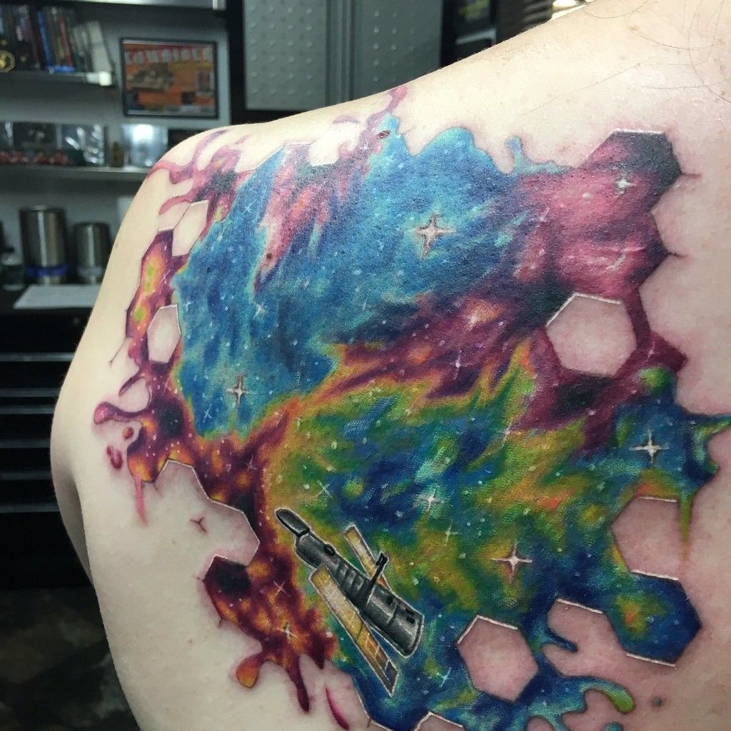 A tattoo on a person's arm showing a patch of the cosmos in different colors: mostly blue and dark pink, with smatterings of yellow and green along with white stars. At the bottom of the patch of universe is the Hubble Space Telescope.