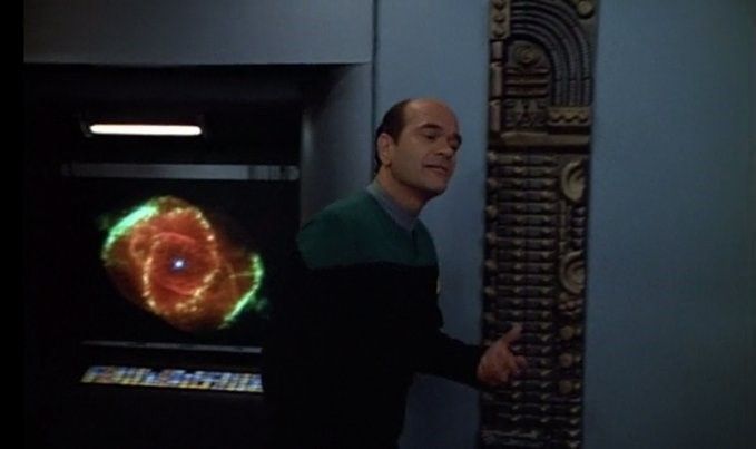 A screenshot from a Star Trek episode, which shows a male character in the center of the screen and, behind him on the left, a colorful Hubble image of a supernova explosion.