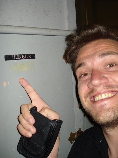 A smiling man on the right side of the image points with his right hand to a gray locker with the word "Hubble" on it in black on the left side of the picture.