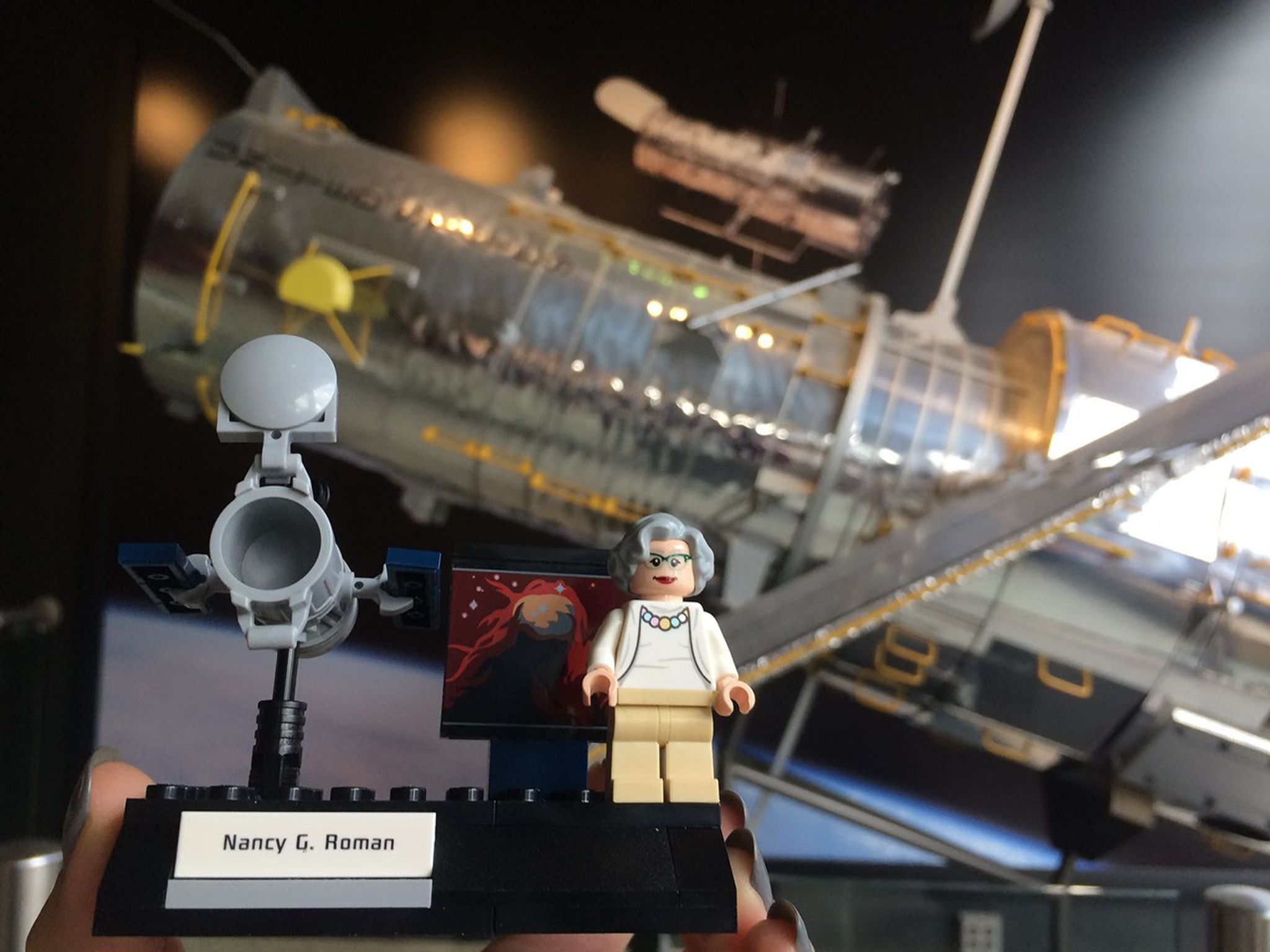 A person holds up a small Lego model of Nancy Grace Roman next to a model of the Hubble Space Telescope with a Hubble image behind her. In the background is the much-larger model of Hubble at Goddard.