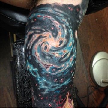 A picture of someone's leg with a large tattoo (covering almost the entire portion of their leg visible in this image) of a spiral galaxy on it. The center of the galaxy is black and light orange, blending into turquoise for each of the spirals with thin streaks of red throughout, all against a black background with smatterings of white-and-red stars.