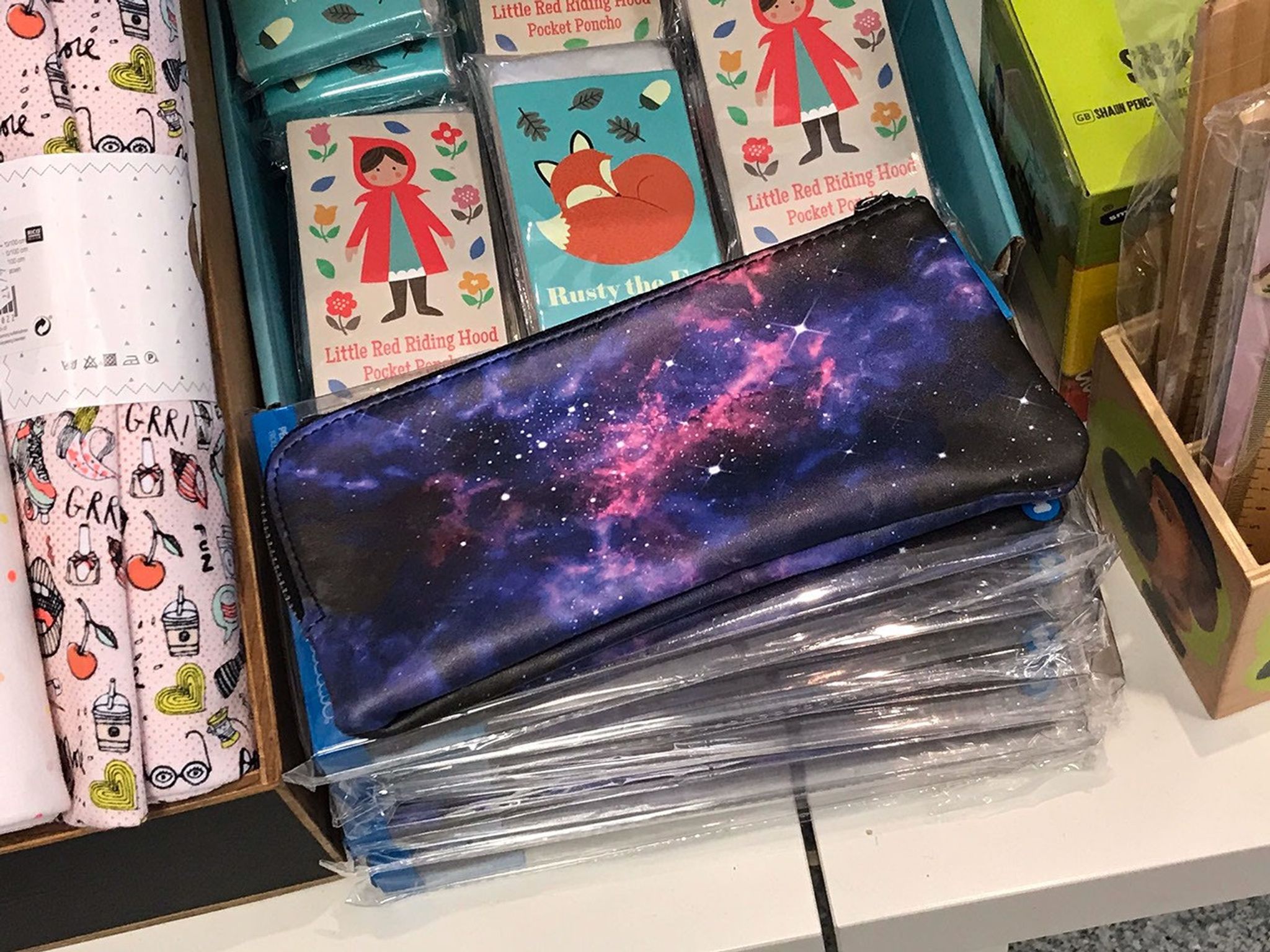 Sitting between various other colorful products on the left, right, and upper center, at the center of this image is a space-themed pencil case, with a nebula of pink and bluish-purple on a black background scattered with many white stars.