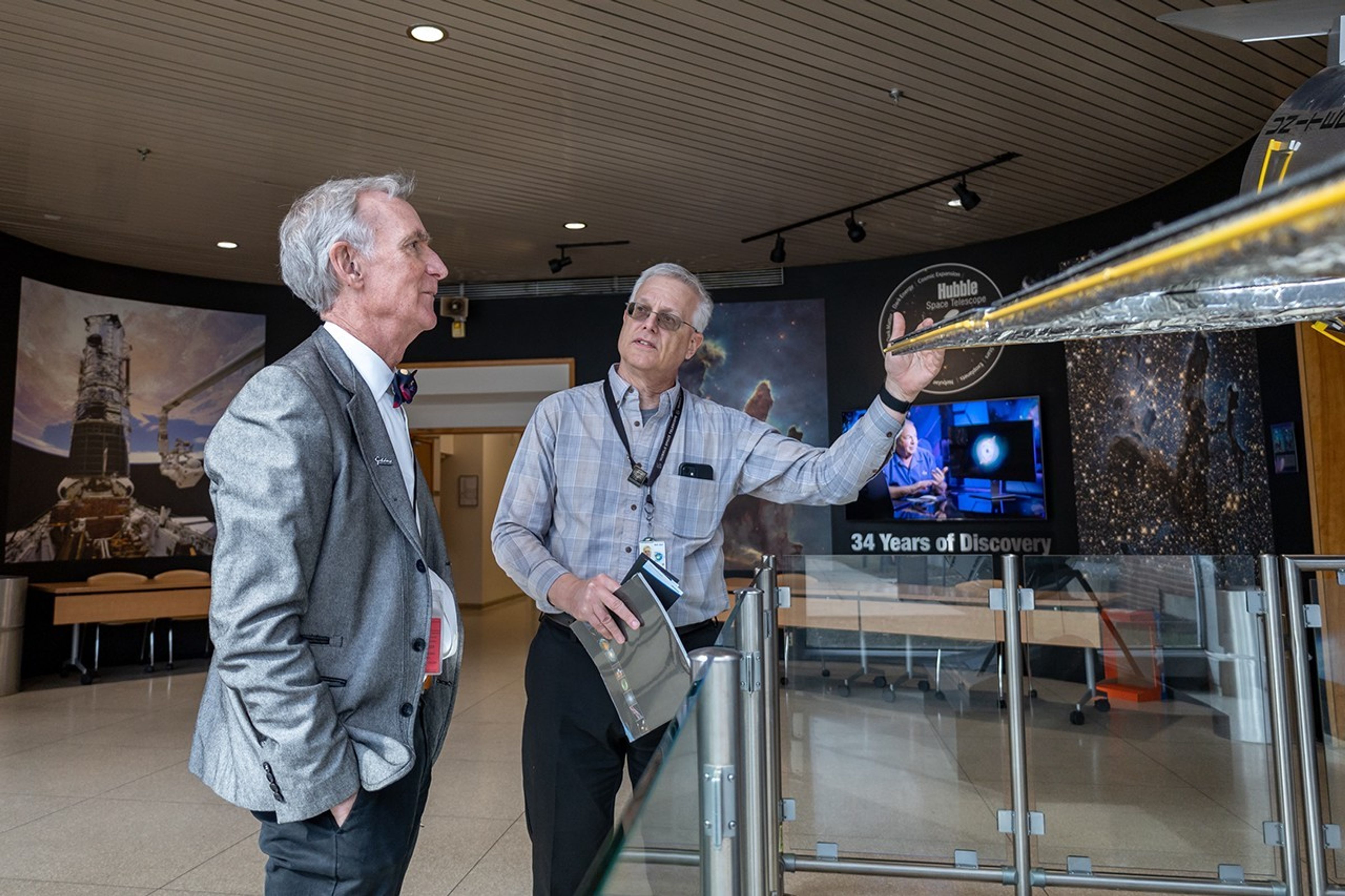 Science communicator and television presenter Bill Nye examines a model of the Hubble Space Telescope at Goddard Space Flight Center. Hubble deputy director Jim Jeletic gestures to the model. Large Hubble images are visible on the walls in the background,.