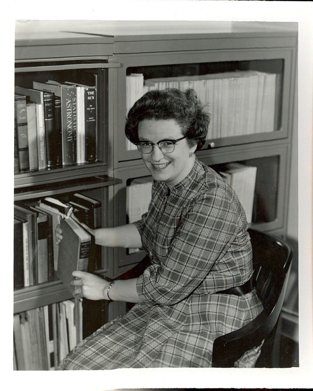 A portrait of Dr. Nancy Grace Roman from NASA's Historical Reference Collection, holding a book while sitting next to a shelf of many more books